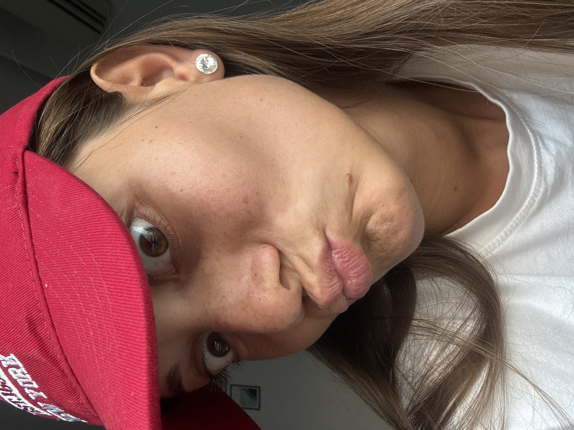 A close-up, angled selfie shows a woman with brown hair and eyes. She is wearing a red baseball cap, which partially obscures her face. She has an earring in her left ear. She is wearing a white shirt. The background is blurry and indistinct. The location is not discernible from the image.