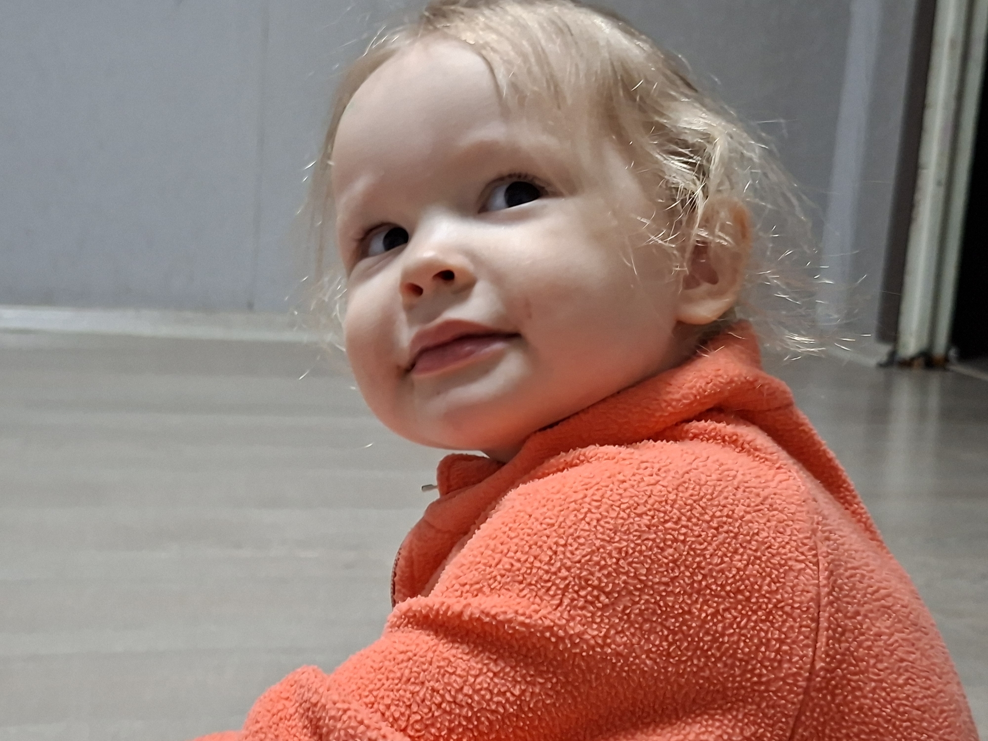 A toddler with fair skin, light blonde hair, and an orange fleece jacket is seen indoors, possibly on a floor, against a backdrop of a light-colored wall and floor. The location is Unknown, Unknown. The child is looking slightly to the left with a small smile. The texture of the fleece jacket is visible, as are the strands of the child's hair.