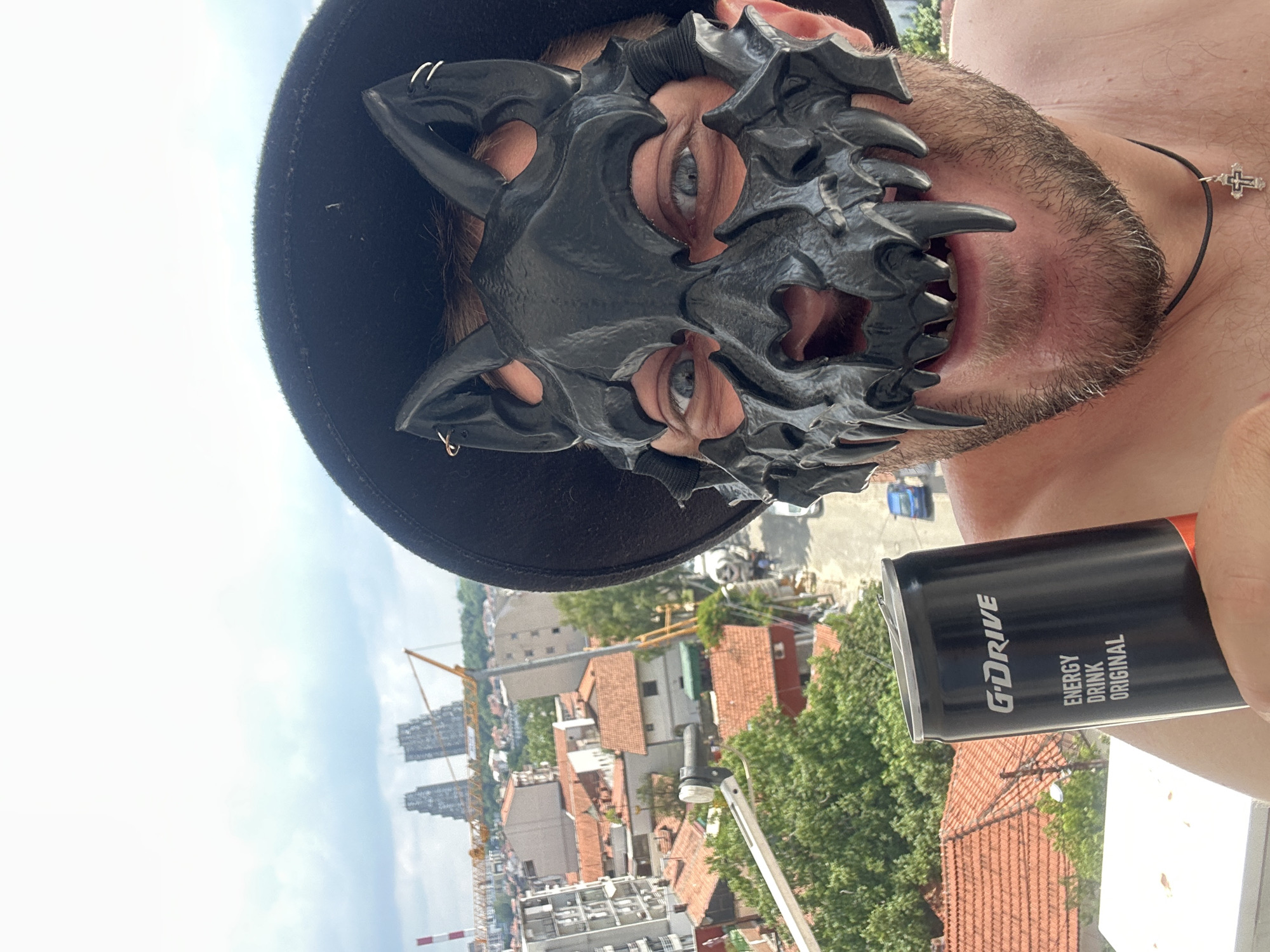 This media file captures a close-up, low-angle shot of a person wearing a black, dragon-like mask and a black hat. The mask covers most of their face, leaving only their eyes and mouth visible. Their eyes are wide and appear to be light blue. The person's mouth is open, revealing their teeth, which are sharp and pointed, mimicking the mask's design. They have a light stubble on their chin and neck, and are wearing a thin black cord necklace with a small silver cross pendant.
The person is holding a black can of "G-DRIVE ENERGY DRINK ORIGINAL" in their right hand, which is partially visible in the foreground.
The background suggests an outdoor urban setting, likely a balcony or rooftop, overlooking a cityscape. The view includes several buildings with red-tiled roofs, dense green trees, and construction cranes in the distance. The sky is bright and appears to be a clear day, indicating it's likely daytime. The orientation of the image is rotated, making the cityscape appear vertical. The overall mood is somewhat mysterious and edgy due to the mask and the intense gaze of the person. The location context points to Zvezdara Urban Municipality, Serbia.