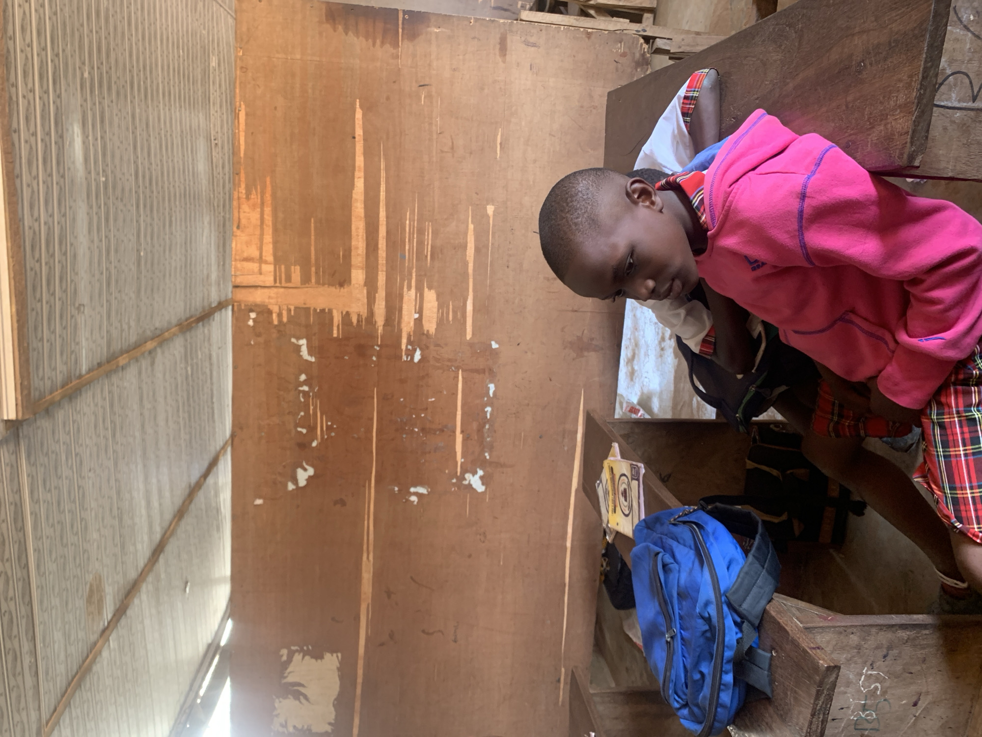 The photograph depicts a child indoors, likely in Ukwu-Abwa, Nigeria, given the user's request. The child wears a pink hooded sweatshirt and a red plaid skirt or shorts. A blue backpack rests on a wooden surface, possibly a desk or a shelf. The interior features unfinished wooden walls with visible markings and wear. The room appears cluttered, with additional objects and structures partially visible. The lighting suggests an indoor environment with natural light filtering through an opening.