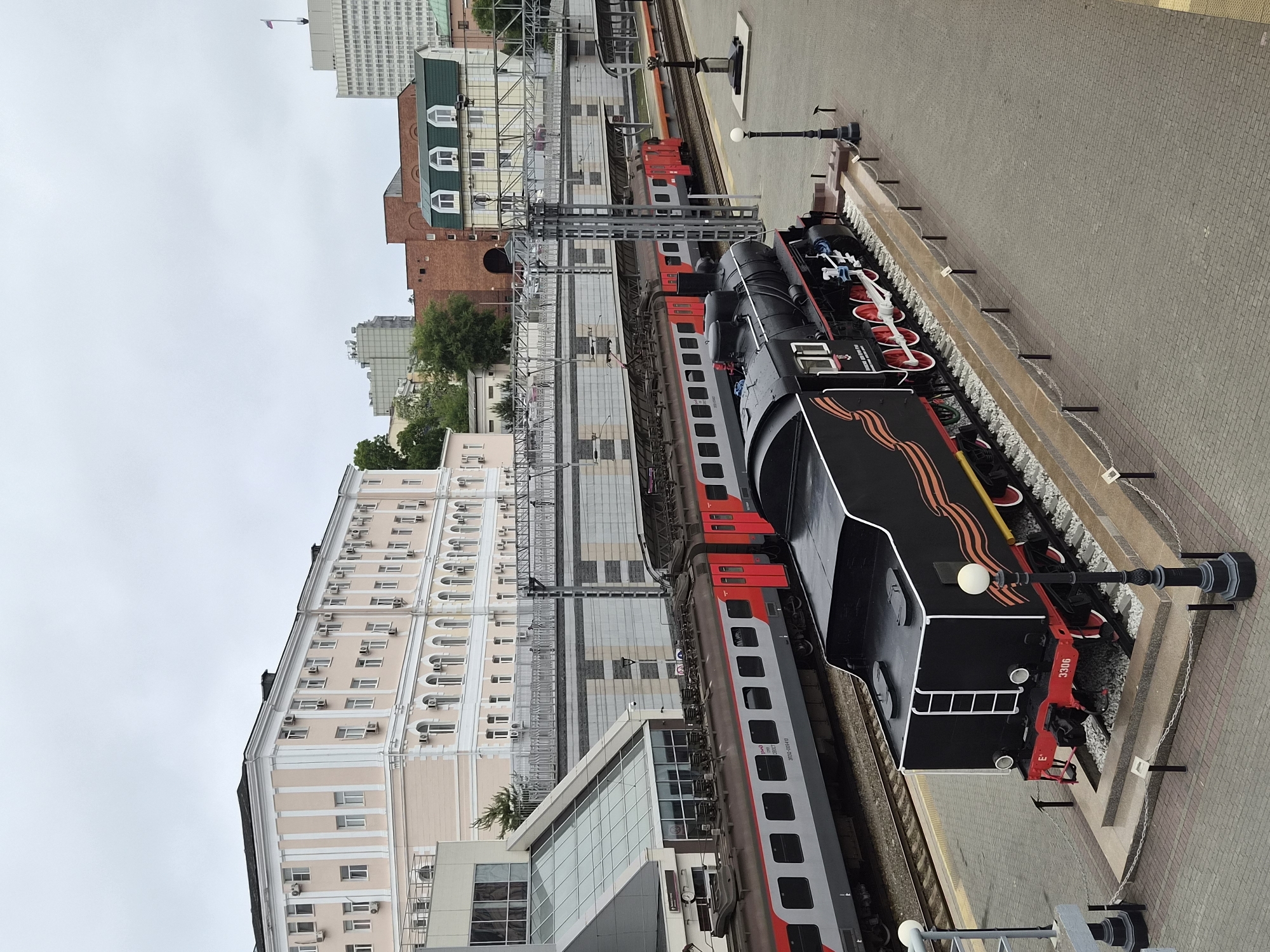 The image shows a railway station in Vladivostok, Russia, on an overcast day. In the foreground, a black steam locomotive, number 3306, is on display. It is decorated with orange and black ribbons. Behind the steam locomotive, a modern red and grey passenger train is visible on the tracks. The station buildings are visible in the background, including a light pink building with white trim and numerous windows, and a red brick building with green-gabled roofs. The scene appears to be a public area, possibly a museum or monument.