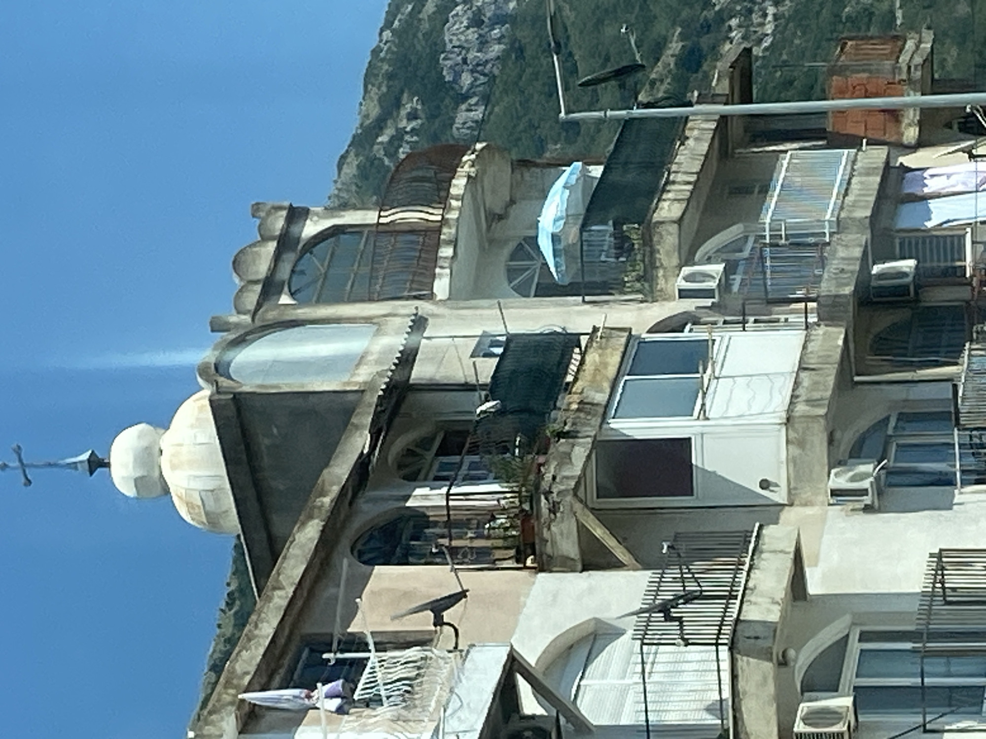 The image shows a multi-story building with a domed structure and a cross on top, located in Mišići, Montenegro. The building appears to be made of concrete and has several balconies with metal railings. Some balconies have plants, and one has a blue umbrella. There are windows and air conditioning units visible on the building's facade. The building is situated on a hillside, with a green, mountainous landscape in the background. The sky is clear and blue. There are no people visible in the image.