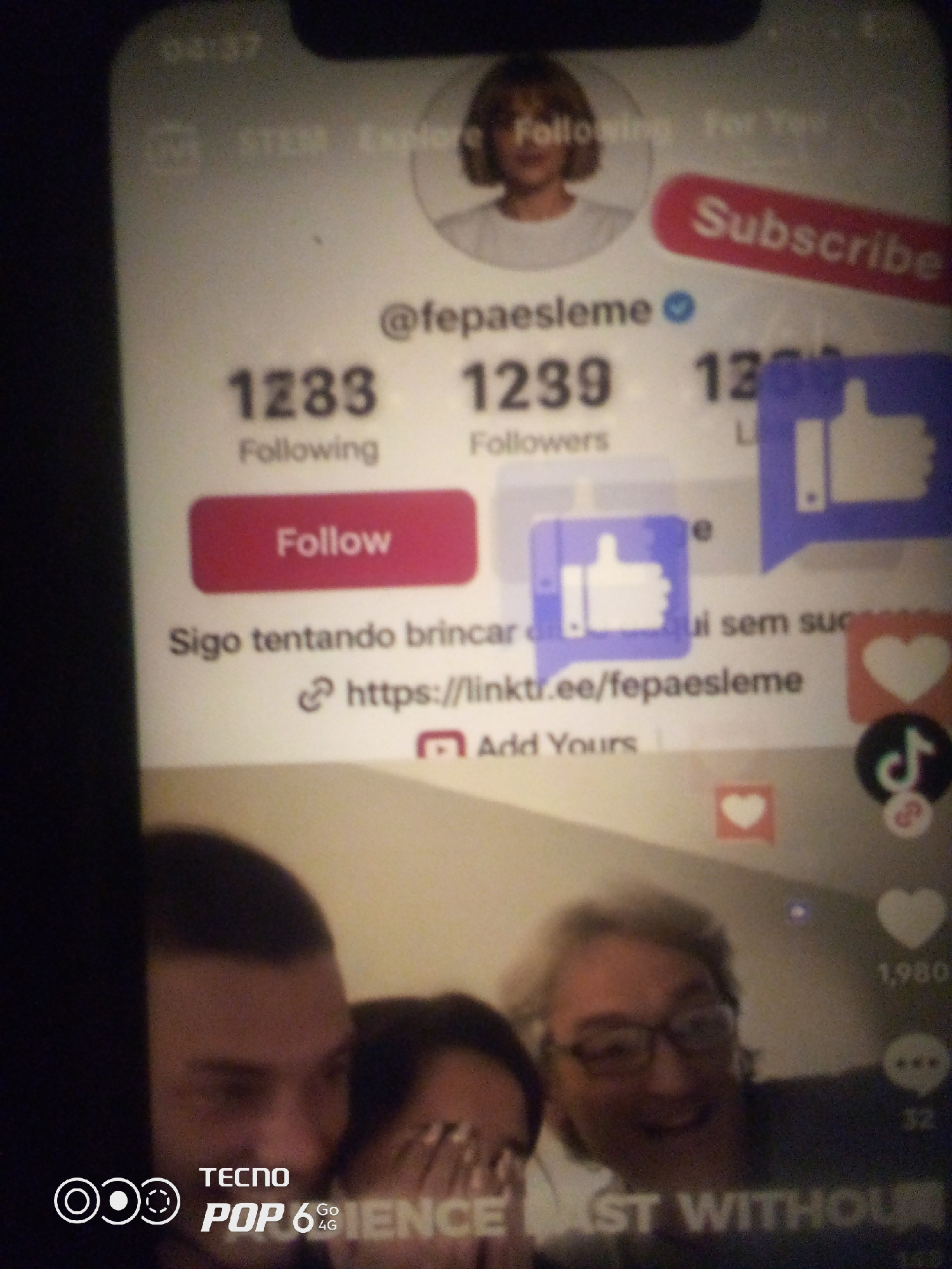 The image is a low-resolution photograph of a smartphone screen displaying a social media profile. The profile belongs to a user with the handle "@fepaesleme". The screen shows follower and following counts: 1233 following and 1239 followers. A "Follow" button is visible, as is a "Subscribe" button. Text below the follower count reads "Sigo tentando brincar aqui sem sucesso" followed by a linktr.ee URL. Various social media interaction icons, including hearts and thumbs-up, are present. A small TikTok logo is also visible. The background of the phone screen displays a profile picture of a person. The bottom portion of the image shows reflections of people and a Tecno Pop 6 4G logo. The image quality is poor, with blurriness and glare. There is no information in the image to suggest it is located in Howayi, Nigeria.