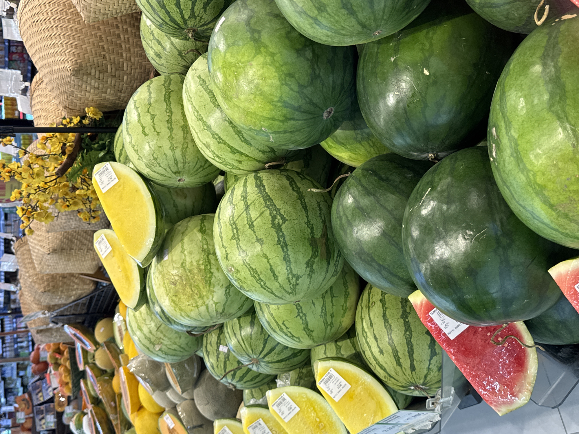 A close-up, eye-level, indoor shot captures a display of fresh fruits at a market in North Kuta, Indonesia. The foreground is dominated by numerous watermelons, varying in size and shade of green, with distinct dark green stripes on some. Several slices of yellow watermelon, showing their bright yellow flesh, are arranged prominently, some with price tags attached. A single slice of red watermelon, revealing its juicy red interior, is also visible. In the background, woven baskets and more fruit, including what appear to be cantaloupes and papayas, are stacked. Yellow flowers add a touch of color to the scene, and the overall lighting suggests a well-lit retail environment, likely a supermarket or a fruit stand.