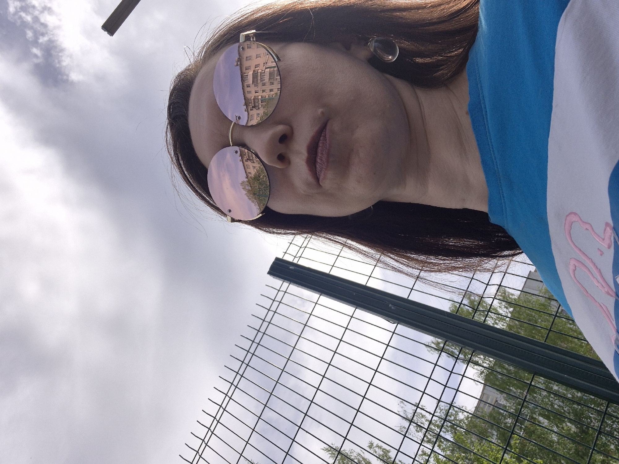 A low-angle selfie shows a woman with long brown hair and light skin. She is wearing round sunglasses that reflect a building with multiple windows on the upper lens and trees and a cloudy sky on the lower lens. She also has a large, clear earring. She is wearing a blue shirt with a white stripe and a pink design. In the background is a green wire fence, trees, a building, and a cloudy sky. The location is Unknown, Unknown.