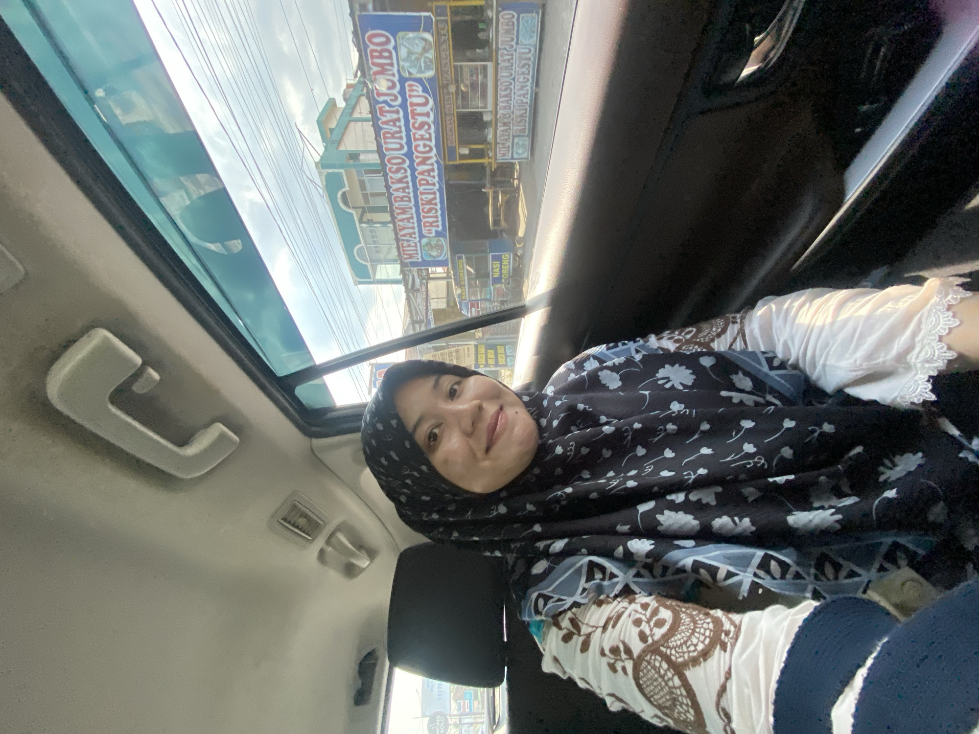 A woman wearing a black and white hijab and a white and brown patterned shirt is sitting in the back seat of a car. She is smiling and looking at the camera. Outside the car window, a street scene is visible with a restaurant named "MIE AYAM BAKSO URAT JUMBO 'RISKI PANGESTU'". The restaurant is located in Banjarbaru, Indonesia. The image is taken during the day, with the sun shining.