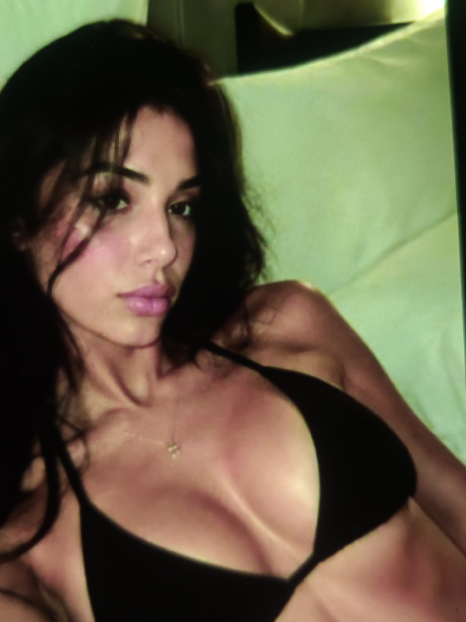 The image shows a close-up of a woman in a black bikini, lying down. She has dark hair and is looking to the side. The background appears to be a bed with a light green headboard. There are no discernible elements in the image that place it in Athens, Greece.