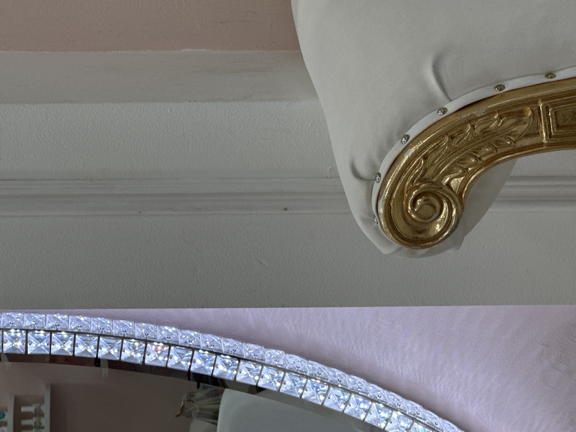 The image presents a close-up, partially obscured view of interior elements. In the upper right portion, a decorative gold-colored element, possibly part of a chair or other furniture, is visible with some studs. The backdrop is a white wall with some texture.
In the lower portion, a fragment of a semi-circular object is visible. It has what appears to be a series of small, square, clear crystals or embellishments along its edge, which could be part of a mirror or decorative lighting fixture. The background behind the semi-circular object transitions to a pink hue.
Given the lack of discernible landmarks or contextual clues, pinpointing the exact location in Sofia, Bulgaria, or any specific place is impossible. No people, interactions, or actions are captured in the frame. The image focuses solely on decorative details.