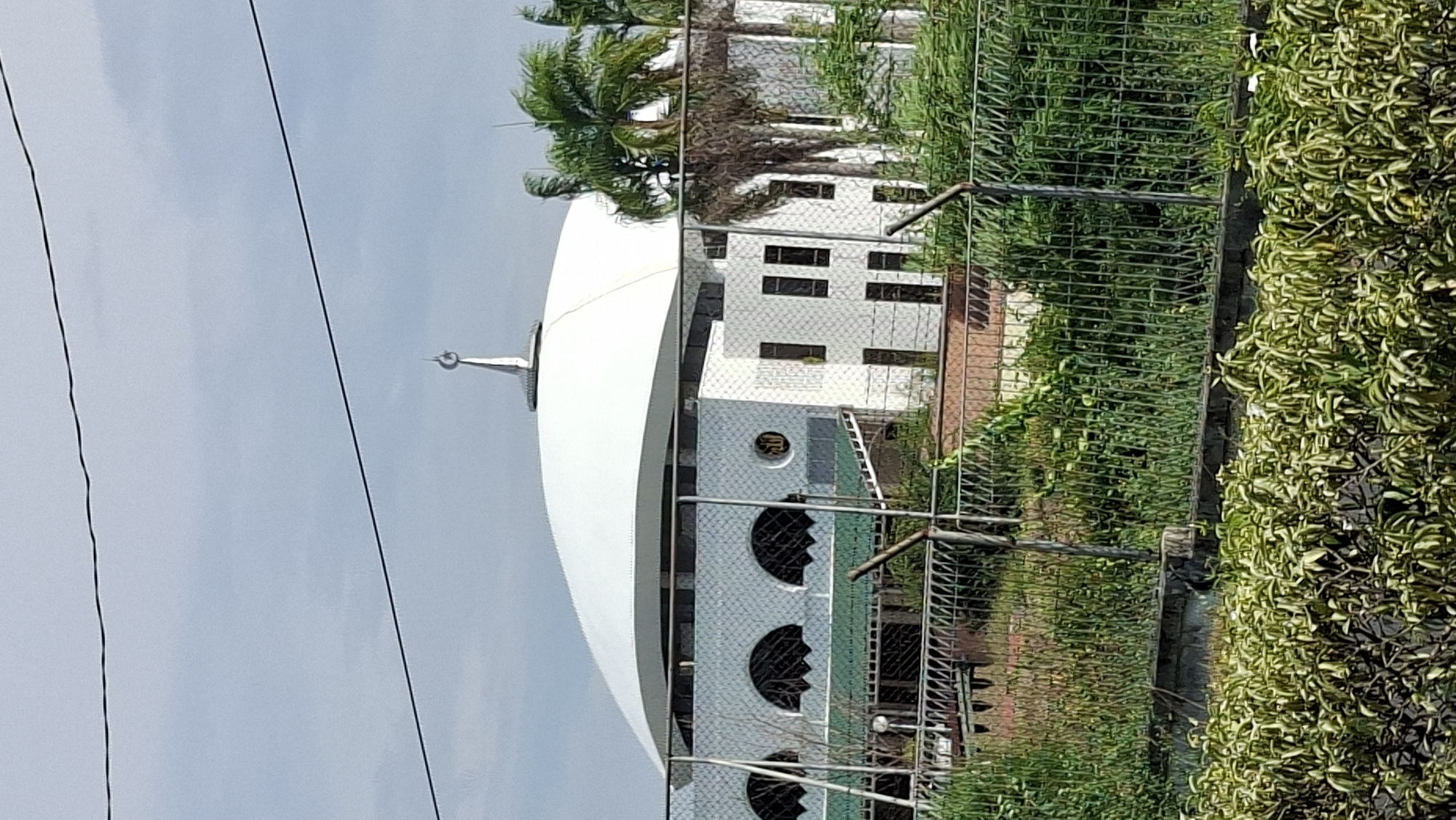 The image shows a mosque with a large white dome and a crescent moon finial, located in Lhokseumawe, Indonesia. The mosque features white walls with patterned tiles and arched windows. In the foreground, a metal fence and lush green vegetation obscure part of the view. Palm trees are visible behind the mosque. The sky is overcast.