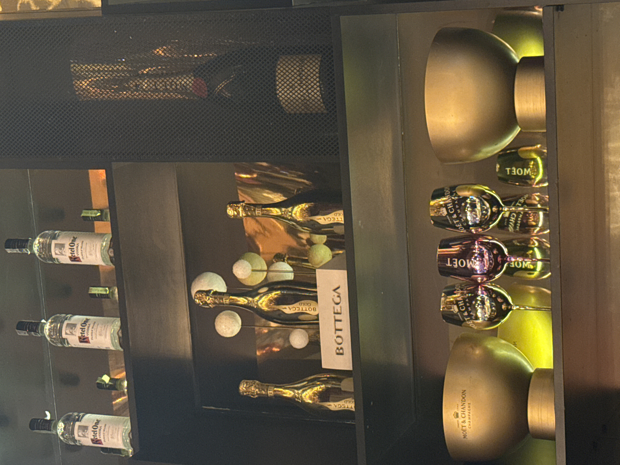 This media file captures a close-up of a well-stocked display shelf or bar cabinet, showcasing various premium alcoholic beverages. The scene is brightly lit, causing reflections on bottles and glassware, suggesting an indoor setting like a bar, store, or restaurant.
On the left side of the display, three bottles of clear liquor are neatly arranged, identified by their labels as "Ketel One VODKA."
The central section features multiple gold-colored bottles, prominently labeled "BOTTEGA GOLD." These bottles are illuminated, creating a strong metallic sheen, and are accompanied by several small, light-colored decorative spheres or orbs. A white sign with "BOTTEGA" written vertically is also visible in this section.
The right side of the display holds items related to "MOET & CHANDON CHAMPAGNE." This includes at least two gold-colored champagne flutes, each bearing the "MOET & CHANDON" logo, and two large, gold-toned ice buckets or coolers with "MOET & CHANDON CHAMPAGNE" inscribed on them. Additional reflections suggest other bottles or glasses in this section.
The top portion of the image is partially obscured by a dark, mesh-like panel, behind which some indistinct bottles or objects can be glimpsed. The shelves themselves appear to be made of dark material, possibly wood or metal, providing a contrast to the gleaming bottles and glassware. No human subjects or activities are visible; the focus is entirely on the presented products. The lighting creates a sense of luxury and highlights the brands on display.