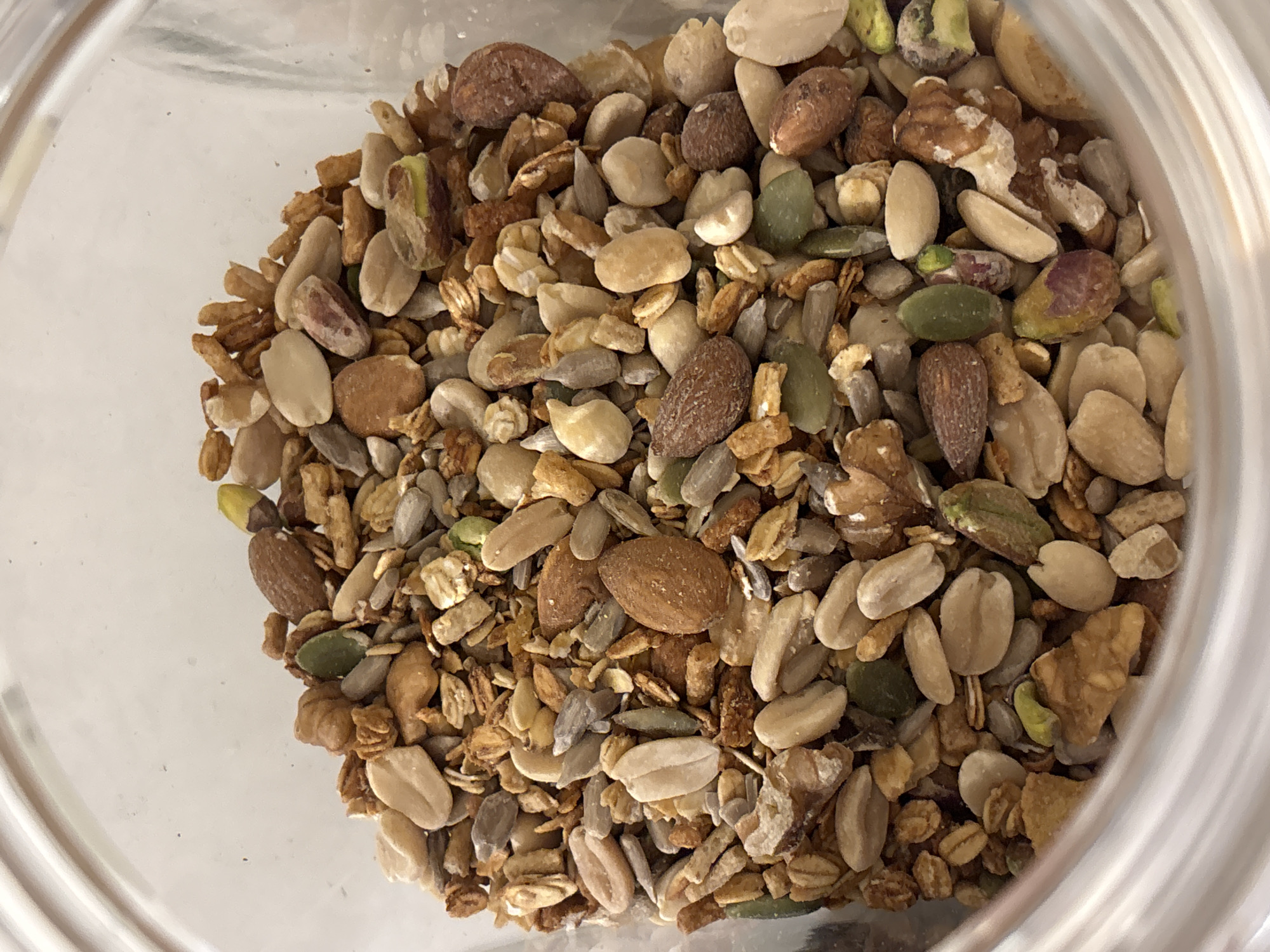 The image provides an overhead view looking down into a clear glass jar filled with a diverse mixture of nuts, seeds, and what appears to be granola. The contents occupy the majority of the frame, showing a rich tapestry of textures and natural earthy colors, predominantly browns, beiges, and greens.
Individual components of the mix are clearly visible. There are numerous whole and split almonds, characterized by their light brown, elongated oval shape. Scattered among these are dark green, flattened pumpkin seeds, as well as pale beige, elongated sunflower seeds. Split peanuts, with their characteristic pale color and shape, are also discernible. Small, irregularly shaped clusters suggest the presence of granola or toasted oats, adding a crunchy texture to the blend. Hints of pistachios, some showing their distinctive green color and others with purplish-brown skins, can also be spotted.
The clear glass container suggests a typical food storage jar or bowl. The lighting is bright and even, allowing for a detailed view of each ingredient. No specific activity is taking place; the image is a still life focused entirely on the food items. There is no visible text within the image. The mix appears dry and ready for consumption, likely as a snack, breakfast topping, or baking ingredient. The scene is set indoors, indicative of a kitchen or pantry environment.
