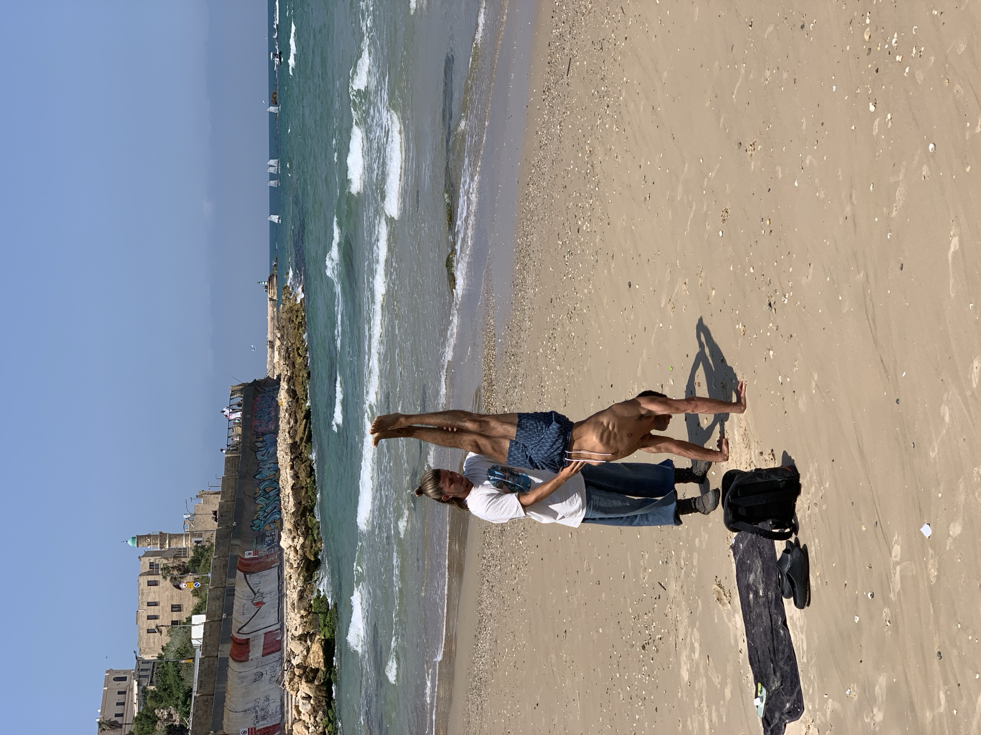 Two men perform an acro-yoga pose on a beach. One man lies on his back on the sand, wearing jeans and a white t-shirt, lifting the other man in a plank position. The second man wears blue patterned shorts and balances in a horizontal position with his hands on the ground. The scene is set on a sandy beach with a cityscape visible in the background, including the Old Jaffa buildings and a seawall. The turquoise water of the Mediterranean Sea laps at the shore, with small boats visible in the distance. Nearby are a black backpack, a black towel, and sandals. The lighting suggests it is daytime, with shadows cast on the sand. This is likely Tel Aviv, Israel.