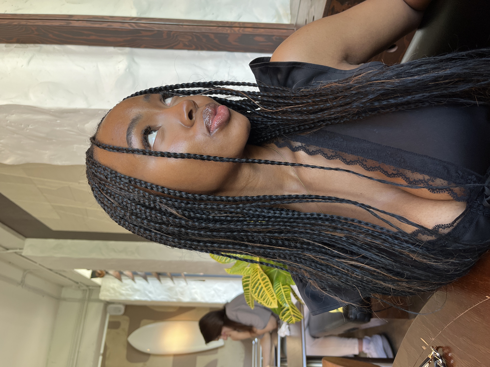 The image shows a medium shot of a young Black woman with braided hair, wearing a black low-cut top with lace trim. She is looking to her right, with her lips slightly parted. She has on lip gloss and her eyes are wide open. She is seated at a wooden table in what appears to be a cafe or restaurant, with a plant in the background and another person blurred in the distance. The lighting suggests an indoor setting with natural light filtering in. There is no visible indication that this scene is located in Kazan, Russia.