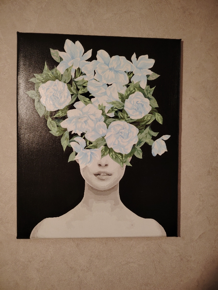 A rectangular canvas painting is affixed to a textured, light gray wall in the city of Марковский, Russia. The painting, oriented vertically, depicts a stylized human figure from the shoulders up against a solid black background. The figure has fair skin, with visible bare shoulders and collarbones. The lower half of the face, including the nose, lips, and chin, is visible. The upper half of the face is obscured by a dense arrangement of light blue and white flowers interspersed with green leaves, which acts as the figure's head or a floral headdress. The canvas itself has visible black edges.