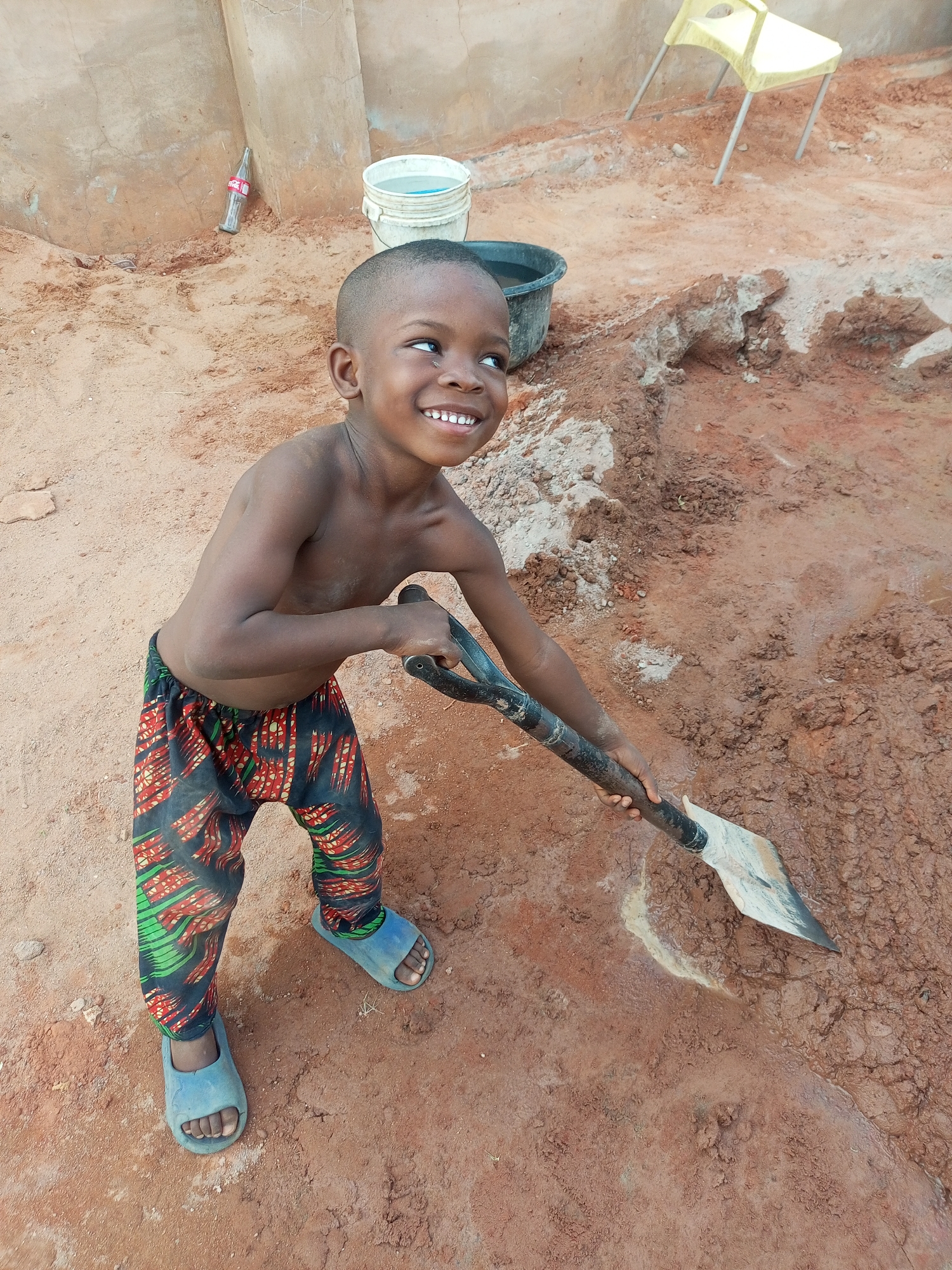 The image depicts a young boy, likely between three and five years old, in an outdoor setting in what appears to be Tempure, Nigeria. He has a short buzz cut, is bare-chested, and wears vibrant patterned pants in green, red, and black, along with light blue slide sandals.
The boy is actively engaged in a task, holding a metal shovel with both hands and pushing its blade into the muddy, red-brown ground. His posture suggests he is either digging or mixing the earth. He is looking upwards and slightly to the right with a wide, joyful smile, revealing his teeth, which conveys happiness and enthusiasm for the activity.
The background features a simple, light-colored concrete wall or structure. To the boy's left, a red glass Coca-Cola bottle is partially visible on the ground, with the "Coca-Cola" text clearly legible on its side. Further back, two buckets are present: one white bucket containing water and a dark bucket. On the right side, a light yellow plastic chair stands against the wall. The ground around the boy is a mix of dry, red soil and darker, wet mud. The scene is brightly lit by daylight, suggesting clear weather.