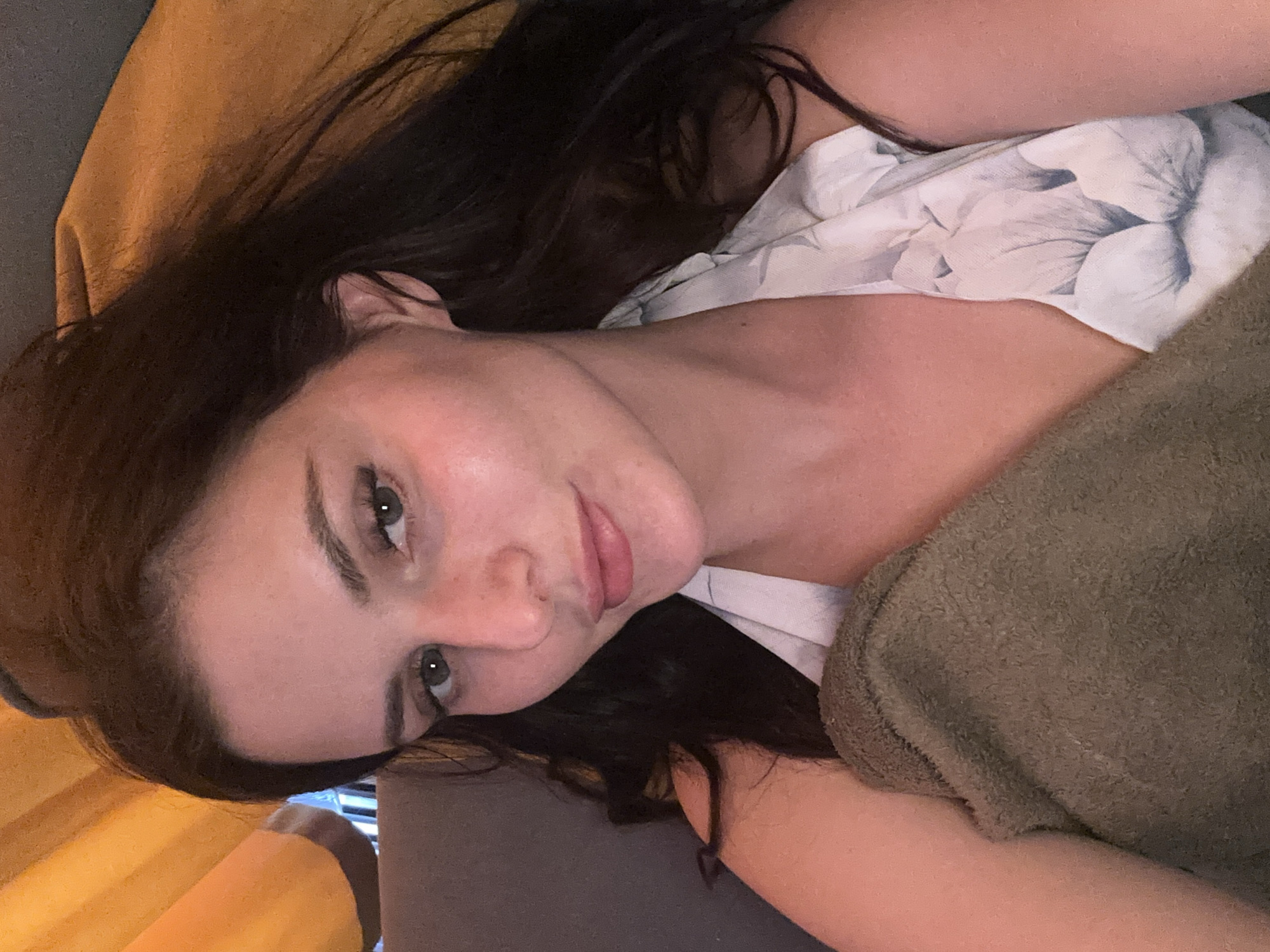 A young woman with long, dark hair is photographed from the chest up, in a close-up selfie. She is wearing a white floral print halter top and is holding a dark olive green towel. Her head is slightly tilted, and she is looking directly at the camera with bright green eyes and a slight smile. Her skin is fair, and she appears to be wearing light makeup. The background is out of focus, with a blurred orange and grey texture that suggests she might be indoors, possibly in a car or on a couch.
