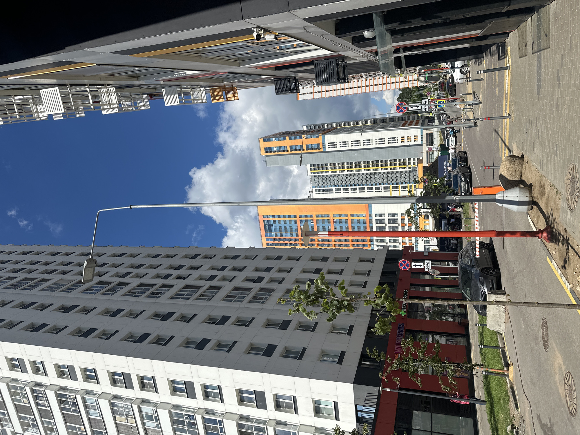 This media file captures a street view in Myakinino, Russia, on a sunny day with scattered clouds. The scene is dominated by modern, colorful apartment buildings with a mix of white, orange, and grey facades. The buildings feature numerous windows and balconies, reflecting the bright sky.
In the foreground, a tall white building with a repetitive pattern of windows stretches upwards. To the right, a street runs alongside the buildings, paved with grey bricks and asphalt. Several cars are parked along the street, including a dark sedan. A prominent red lamppost stands near the center of the frame.
Signs are visible along the street, including a red circular sign with a diagonal bar and a red cross, indicating a prohibition of stopping or parking. Text is visible on one of the buildings, appearing to be in Cyrillic script, with partial words like "РИТМ" and "WILDERMESS."
The image seems to be taken from a street-level perspective, looking up towards the buildings and the sky. The bright sunlight and clear blue sky suggest it's daytime, likely late morning or afternoon. The overall impression is one of a developed, urban residential area.