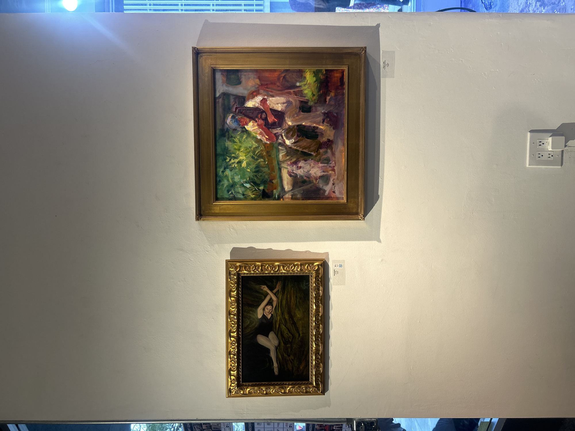 The image shows two paintings hanging on a white wall. The top painting is a square framed oil painting with a depiction of a person reclining or seated in a garden setting. There is a signature at the bottom right corner of the painting. The lower painting is an upright rectangular framed oil painting with a portrait of a dancer. Both paintings are framed in gold-colored, ornate frames. A light shines from above. To the right of the paintings is an electrical outlet. There is a white rectangular label above each painting. Based on other visual cues, the location is likely an art gallery or show in Los Angeles, United States of America.