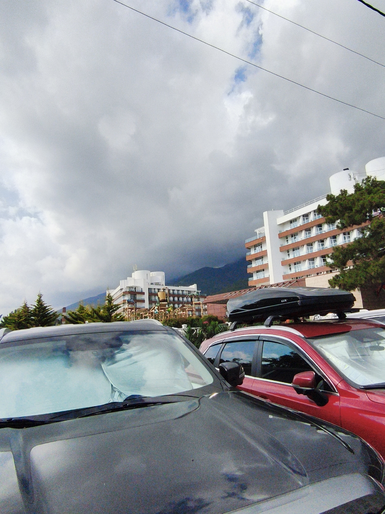 The image captures a cloudy day in Gelendzhik, Russia, with a view of several buildings and vehicles. In the foreground, the hoods of two cars are visible. The car on the left is dark grey, and its windshield is covered by a sunshade. To its right, a red SUV is parked, featuring a black roof box labeled "LUX" mounted on its roof rack. The SUV also has tinted windows.
Behind the cars, a large, modern hotel complex stands out. It's a multi-story building with white and orange accents, featuring numerous balconies with glass railings and patterned facades. To the right of this main building, a taller, cylindrical white structure is visible, possibly part of the same resort. Further in the background, more buildings and trees can be seen. The sky is overcast with dramatic, dark clouds, suggesting an impending storm or recent rainfall, though there are also patches of lighter sky and hints of blue. The overall atmosphere is subdued due to the weather. The presence of pine trees and the architectural style of the buildings suggest a resort town location. No people are visible in the frame.