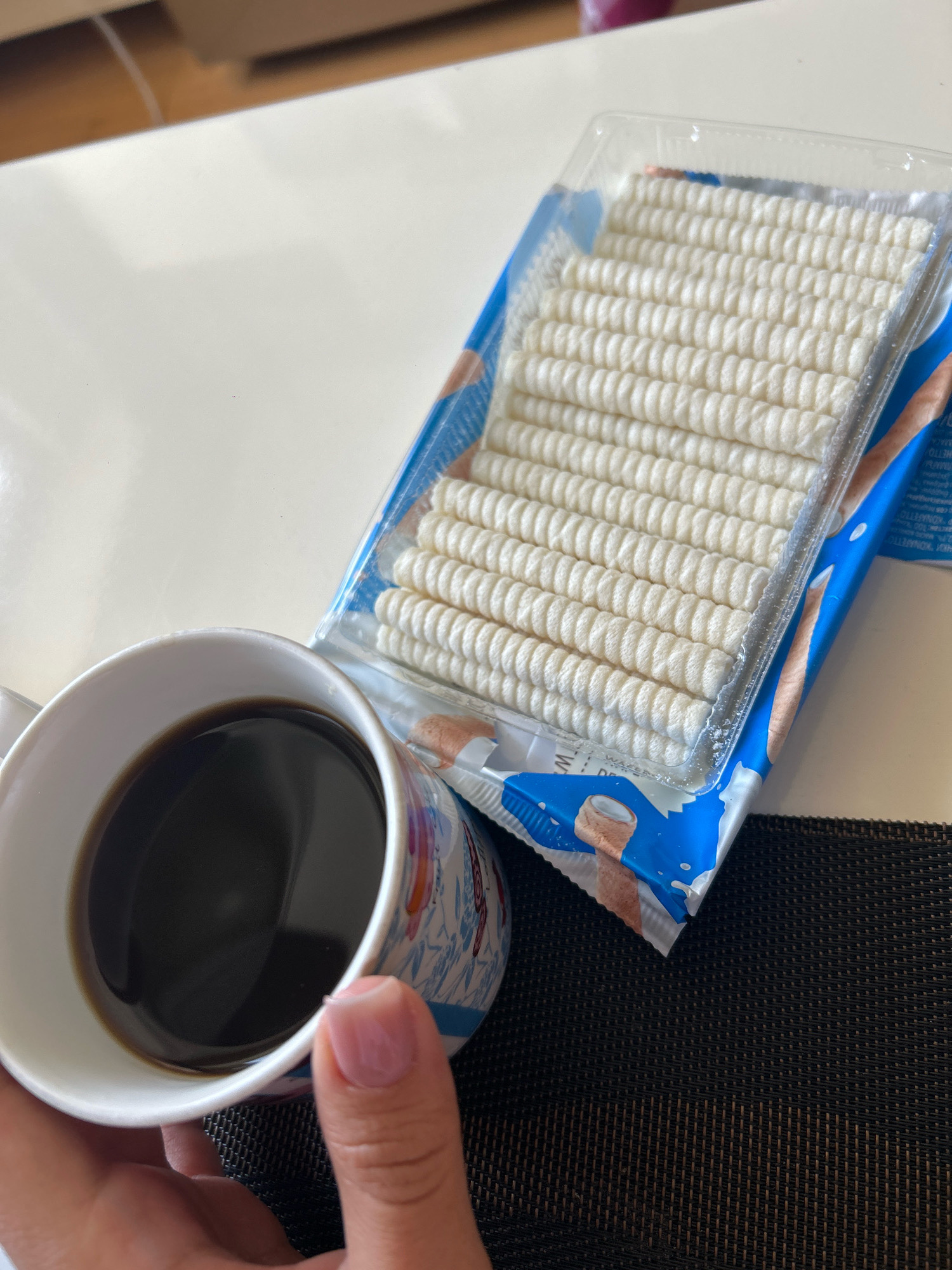 A close-up shot shows a hand holding a white mug filled with dark coffee. Beside the mug is a clear plastic package containing rows of white, cylindrical wafers with a spiral pattern. The package has blue and white branding with imagery of ice cream cones and liquid splashes. The scene is set on a white table, with a dark textured placemat visible underneath the mug and package. The background is blurred, but suggests an indoor setting, possibly a home or cafe. The image does not provide any information about the city of Khyrdalan, Azerbaijan.