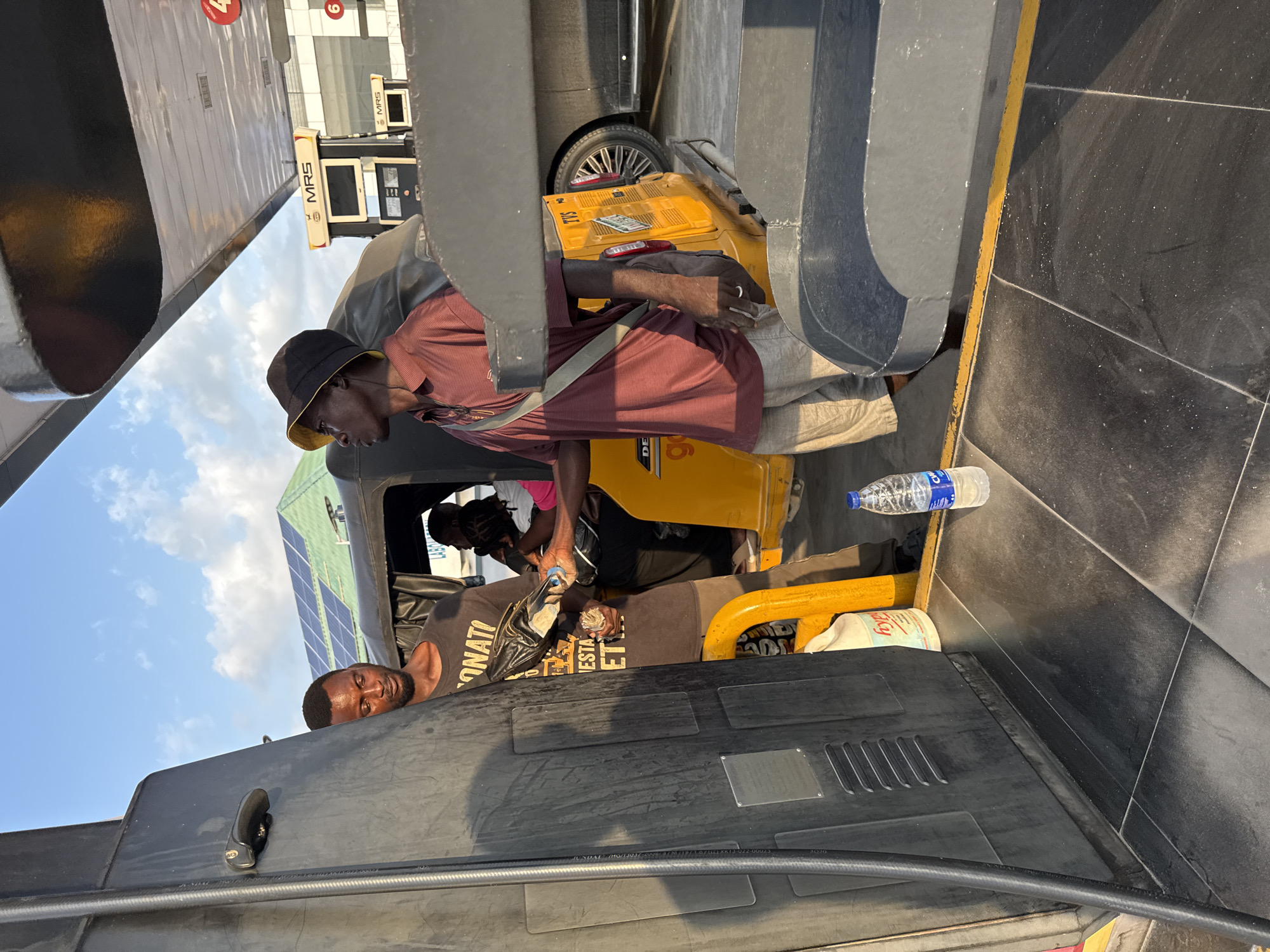 In Ado, Nigeria, at a gas station, two men are visible near a fuel pump. One man, wearing a dark t-shirt with lettering and a black bag in his hand, appears to be interacting with another individual. The second man, dressed in a red and white striped polo shirt and a bucket hat, is looking to his left. Behind them, a yellow motorized tricycle and a dark sedan are parked. The scene takes place under a bright sky with clouds, and solar panels are visible on a building in the background. A plastic bottle of water rests on the dark, tiled ground.