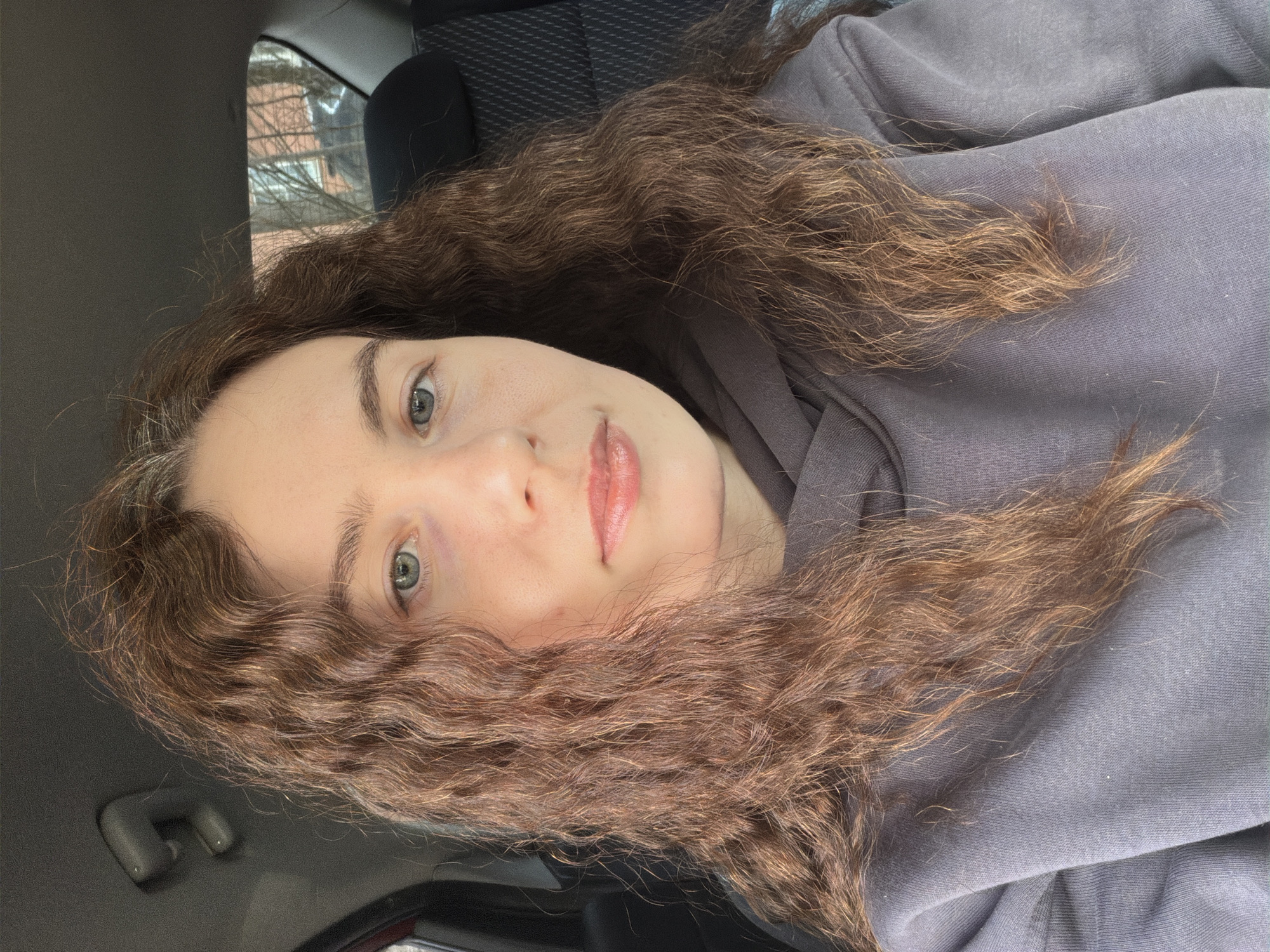 A woman with light skin, green eyes, and brown curly hair is in the passenger seat of a car. She is wearing a grey hoodie. The car's interior has a black headrest and a black and grey textured seat cover. Outside the car window, blurry buildings and bare tree branches are visible. This image does not contain enough information to pinpoint a location in the city of Vsevolozhsk, Russia.