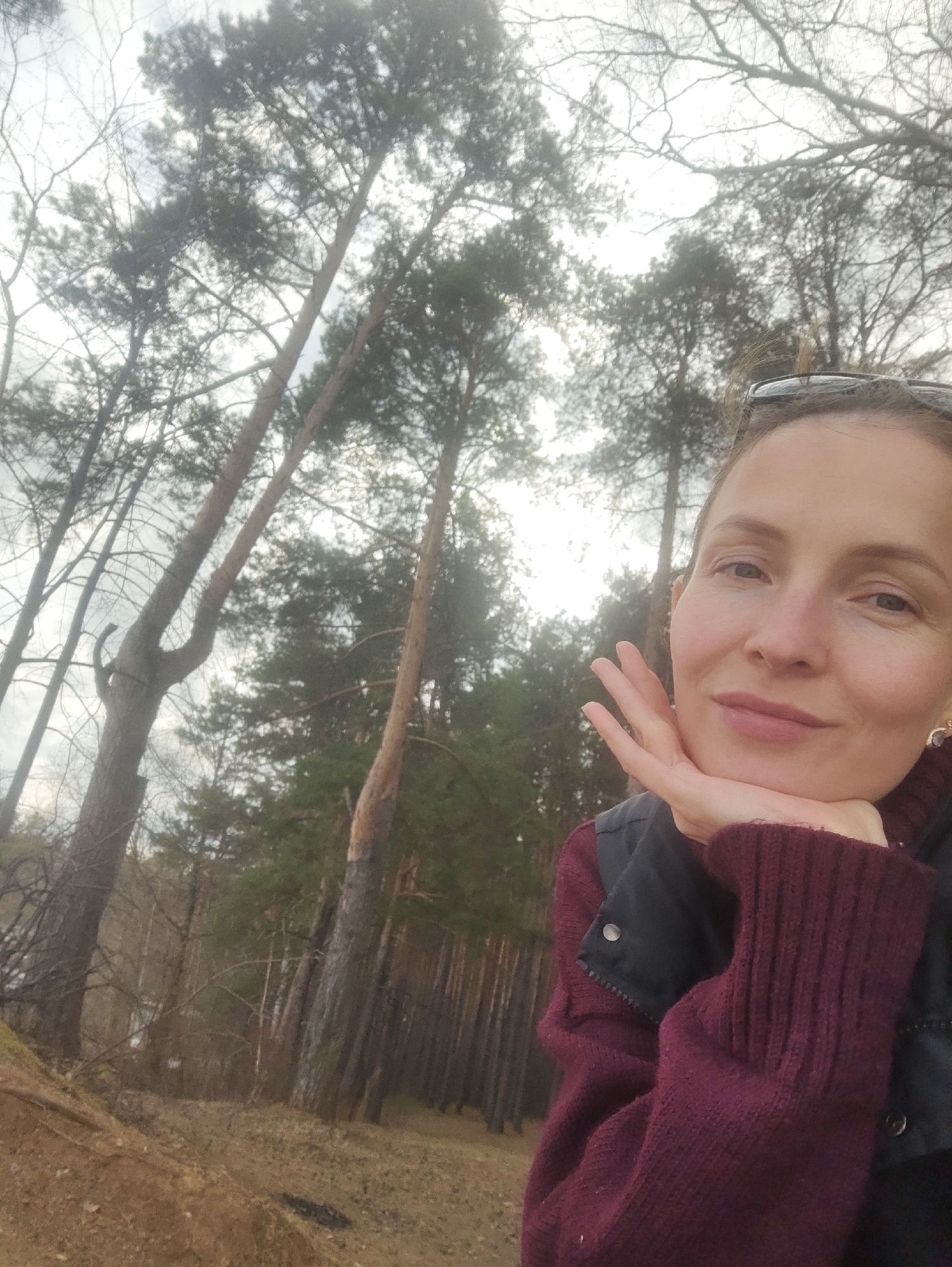 A woman is captured in a close-up selfie, set against the backdrop of a forest in Chaikovsky, Russia. She is looking directly at the camera with a calm, gentle smile, her chin resting lightly on her left hand. Her hair is pulled back, and a pair of dark sunglasses is perched on her head. She is dressed in a dark, possibly olive-green, collared jacket over a warm-looking, chunky-knit burgundy sweater, suggesting cool weather conditions. A small, golden earring is visible on her right ear.
The background reveals a dense wooded area with numerous tall, slender trees. Many are coniferous, likely pines, with dark green foliage concentrated at their tops, while some bare-branched deciduous trees are also present. The sky is overcast and bright, diffusing the light and creating soft illumination without harsh shadows. The ground in the lower portion of the image shows dry, light brown earth and scattered foliage, possibly indicating a season like late autumn or early spring. The woman appears content and relaxed in this natural outdoor setting. No text is visible in the image.