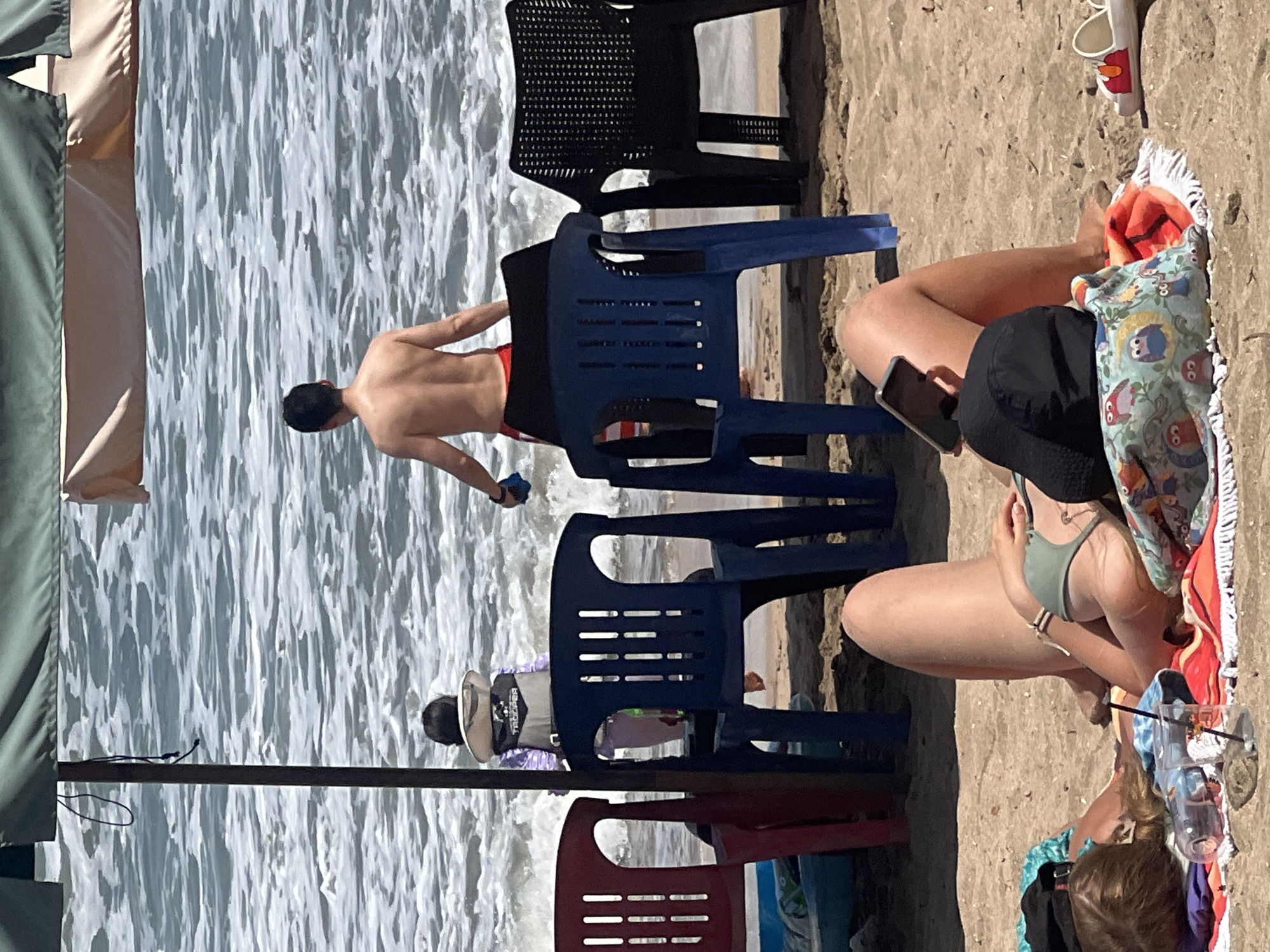 This image captures a sunny day at Kuta Beach in Indonesia. In the foreground, two individuals are relaxing on beach towels on the sand. One person, seen from the torso up, is wearing a green bikini top and holding a smartphone. Another person is lying on their side, with their head resting on a colorful patterned cushion, wearing a black cap. In the middle ground, several plastic chairs are arranged on the sand. Behind the chairs, the ocean is visible with waves breaking on the shore. Two people are in the water: a person wearing a life vest, possibly a child, and an adult male with his back to the camera, wearing red shorts. The scene is set under a large beach umbrella, providing shade.