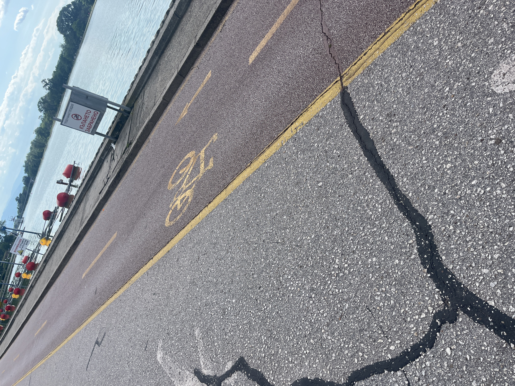 The image shows a bicycle lane in Plovdiv, Bulgaria, next to a body of water, likely the Maritsa River or a canal. The bicycle lane is separated from the adjacent grey asphalt road by a yellow line. A bicycle symbol is painted on the lane. A warning sign is visible near the edge of the water. Red and yellow buoys float in the water. Cracks in the asphalt road have been sealed with black tar. The sky is partly cloudy.