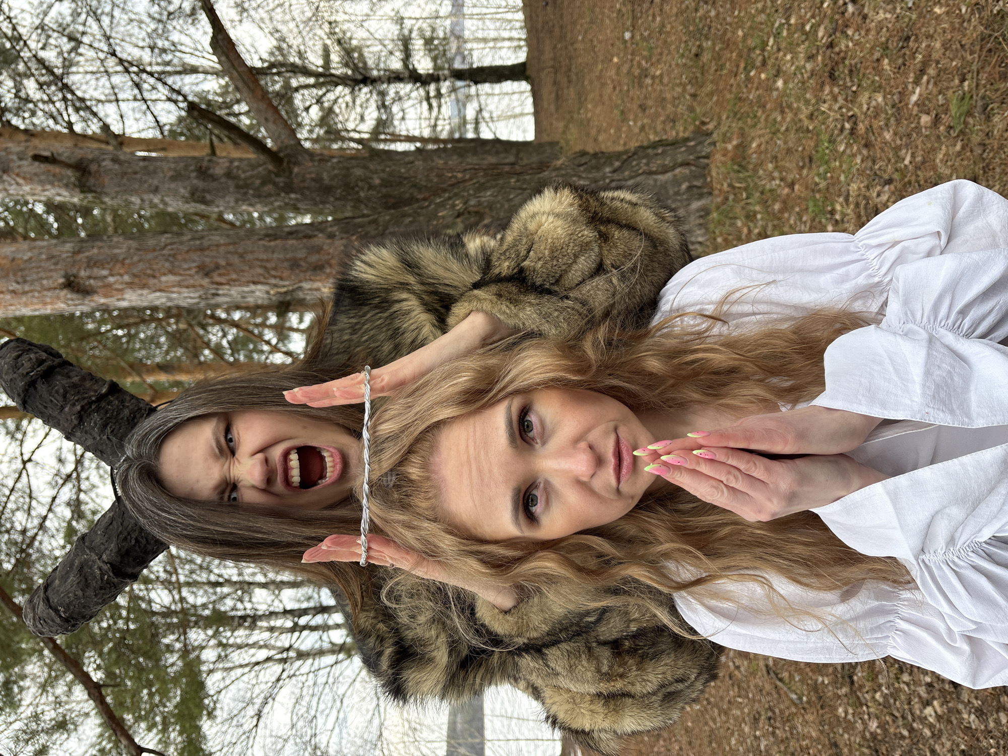 The image shows two women outdoors, seemingly in the city of Chaikovsky, Russia. The background features trees and foliage, suggesting a wooded or park-like area. The woman in the foreground has blonde hair and is wearing a white blouse. She has her hands clasped near her face. Behind her is a second woman wearing a fur coat or shawl. This woman has dark hair with grey streaks, is holding a twisted wire above the blonde woman's head, and is making a facial expression with her mouth open, teeth bared, and eyes wide. A dark, textured, possibly decorative object shaped like a horn is positioned near the second woman's head.