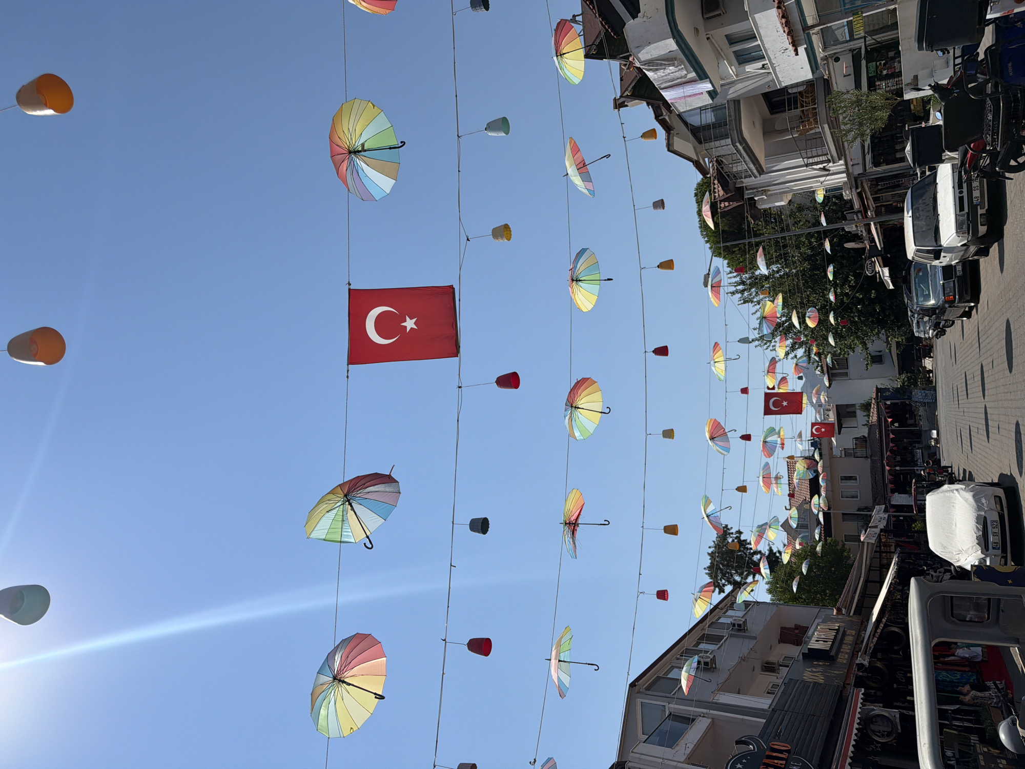 The image shows a street scene in Marmaris, Turkey, adorned with a colorful display of open umbrellas and hanging lights strung across the thoroughfare. The Turkish flag is prominently displayed, hanging from a wire in the center of the frame. Buildings line both sides of the street, with shops and awnings visible on the left. Several cars are parked along the road, and a motorcycle is parked on the right. The sky above is a clear, bright blue. The scene appears to be in an urban environment, likely a commercial area or town center.