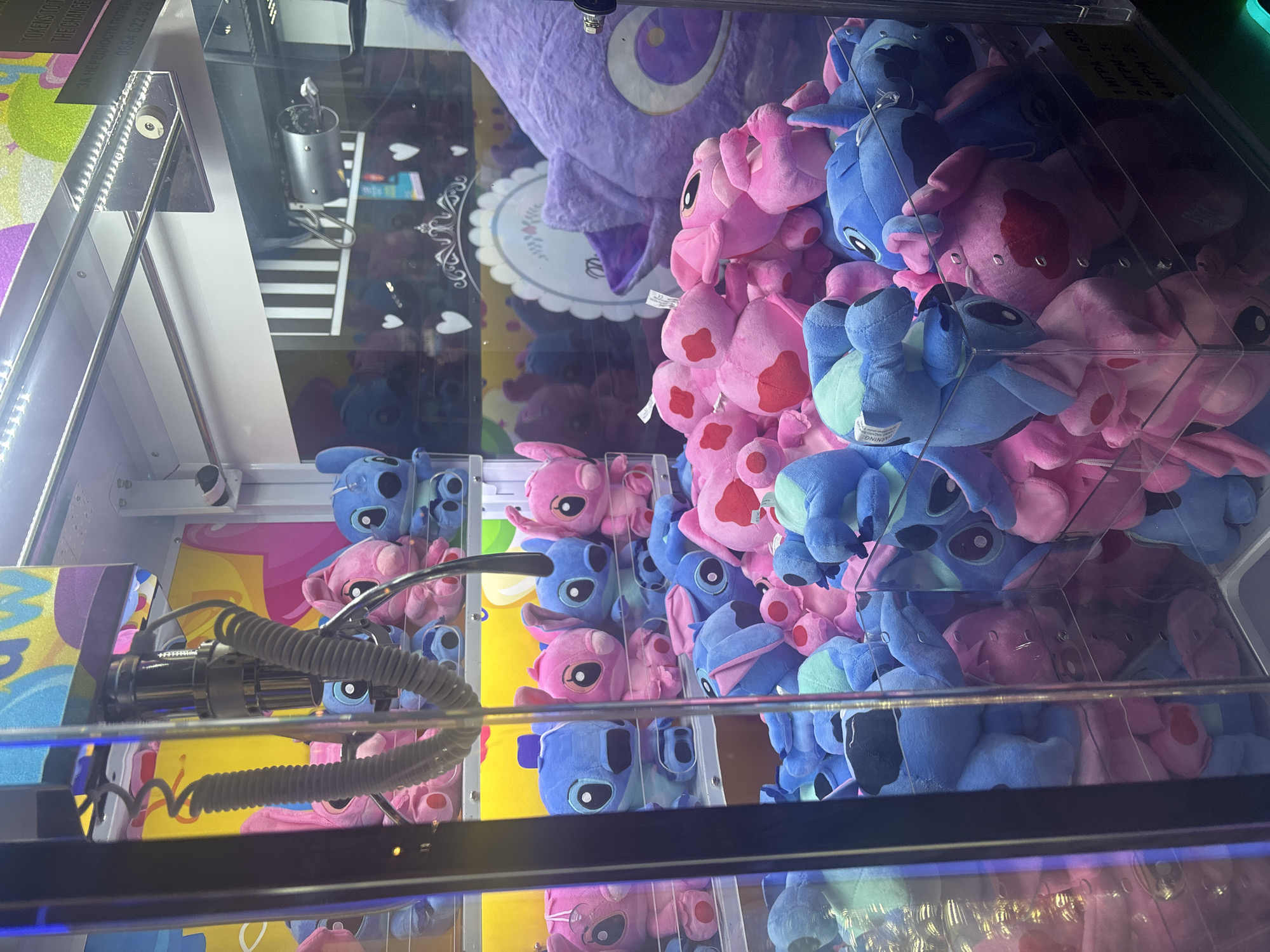 The image shows the interior of a claw machine filled with plush toys, predominantly blue and pink Stitch characters from the Disney movie "Lilo & Stitch", along with a large purple plush toy in the background. The claw mechanism is visible, positioned to potentially grab a toy. Text in Cyrillic is visible, stating price for games and possibly location within Plovdiv, Bulgaria.