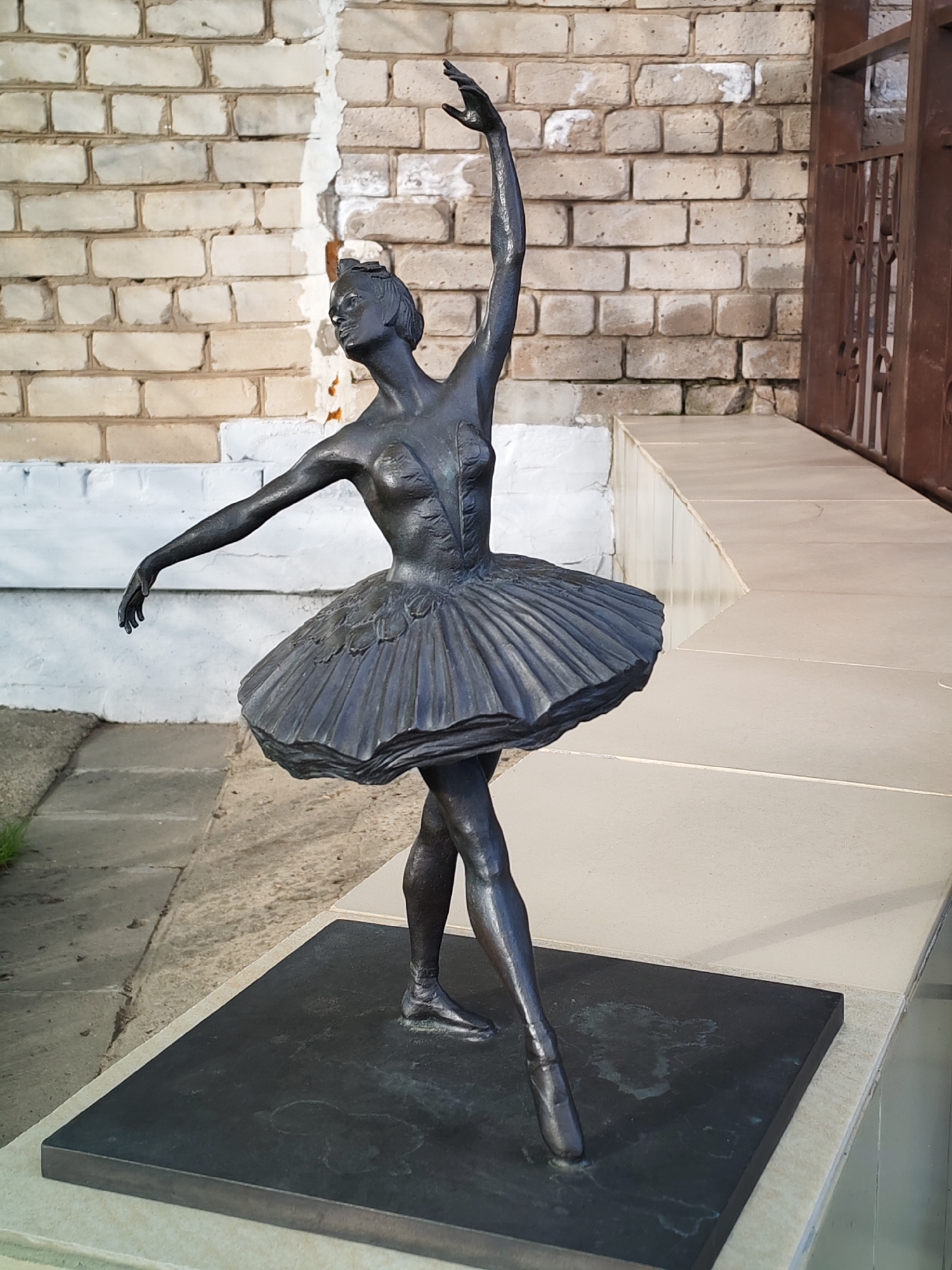 The image displays a graceful, dark bronze or metal statue of a ballerina. The statue depicts a dancer on pointe, with her right leg extended back and her left leg forming the support. Her right arm is raised elegantly above her head, while her left arm is extended to the side, palms facing downwards. She wears a classical ballet costume, including a fitted bodice with sculpted details and a flowing, pleated tutu. Her head is tilted upwards, and her facial features are finely sculpted, conveying a sense of serenity and focus.
The statue rests on a dark, rectangular base, which is placed on a light-colored, paved step or platform. Behind the statue, a textured brick wall with varying shades of light brown and off-white is visible, showing some areas where white paint has chipped away. To the right, a dark brown metal gate or fence with intricate patterns is partially seen. The ground beneath and around the statue's base consists of light-colored paving stones, with some natural ground and sparse grass visible towards the left.
The scene is brightly lit by natural daylight, suggesting a clear day. The detailed sculpting of the ballerina's form and costume is prominent. The setting appears to be an outdoor public area, possibly a park or courtyard, in Chaikovsky, Russia. No visible text is present on the statue or in the background.