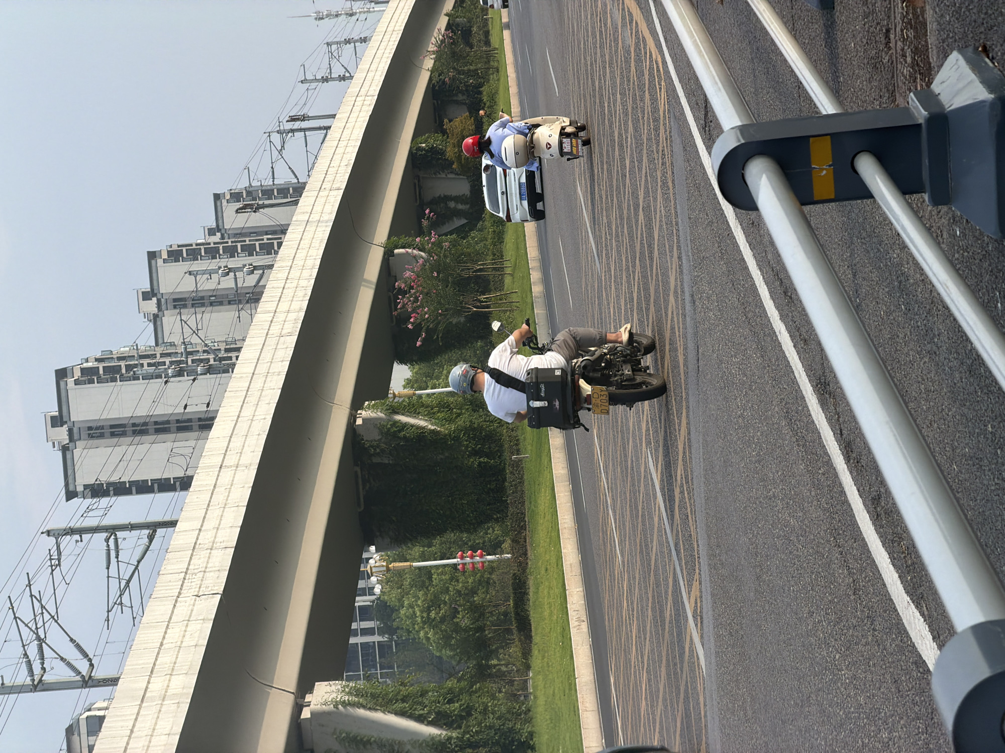 The image captures a street scene in Jiangjin, China, under an elevated overpass, likely for a railway. In the foreground, a gray metal guardrail separates the road from the viewer. The road itself is paved and marked with yellow and white lines, indicating traffic lanes and a pedestrian crossing. Two individuals on scooters are moving away from the camera down the road. The person on the left is riding a white scooter, wearing a white t-shirt, gray pants, and a helmet. They have a black storage box mounted on the back of the scooter. The person on the right is riding a cream-colored scooter, wearing a blue shirt and a red helmet. This scooter also has a white storage box on the back. Further down the road, a white car is visible. In the background, a series of modern, multi-story buildings line the horizon, and lush green trees and vegetation grow along the side of the road and climb up the pillars of the overpass. Overhead, power lines and infrastructure associated with the elevated structure are visible against a hazy sky.