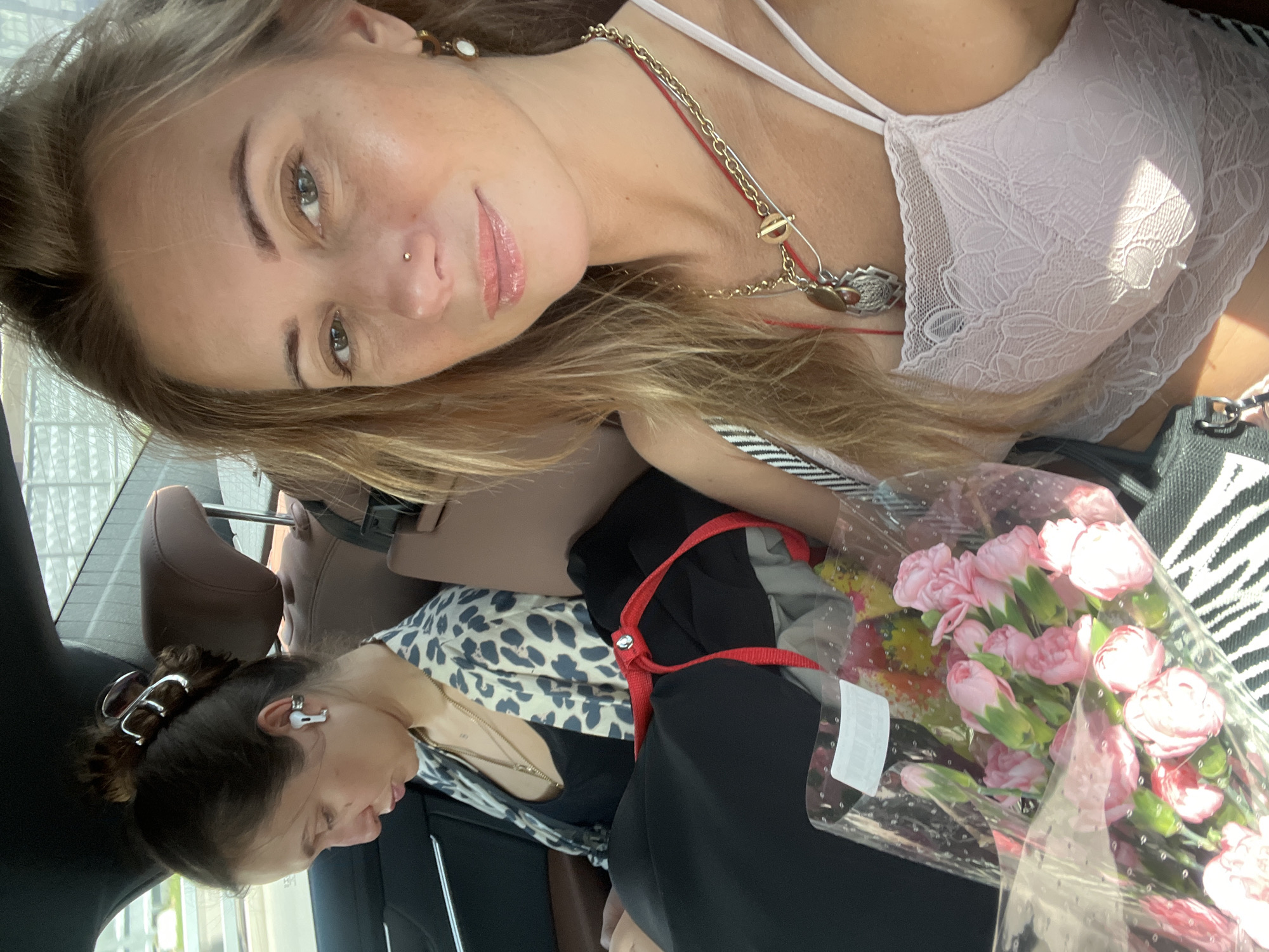 The image captures two women inside a vehicle, likely a car. The woman in the foreground is taking a selfie and is looking directly at the camera. She has long blonde hair, a nose piercing, and is wearing a white lace top and multiple necklaces. A bouquet of pink flowers is visible in her hands. To her left, another woman is seated in the back seat, looking down and to her left. She has dark hair pulled back and is wearing a black top and a leopard-print cardigan. She has earbuds in her ears and is wearing a necklace. The interior of the car appears to be luxurious with brown leather seats. The background suggests they are on a road, possibly in or near Moscow, Russia, given the context. The scene is brightly lit, indicating it is daytime.