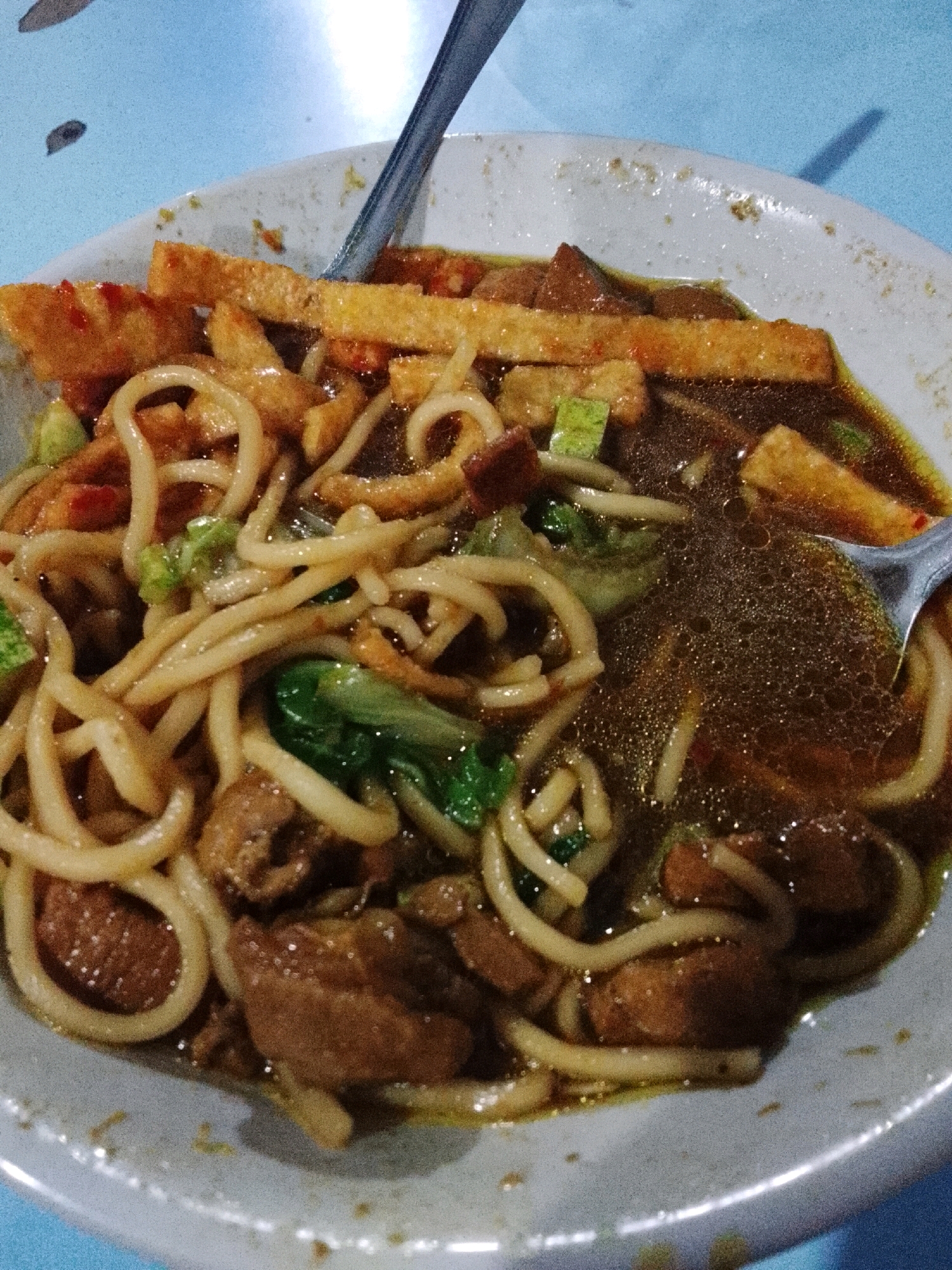 In Padang Dalom, Indonesia, a close-up, overhead shot captures a bowl of noodle soup. The soup, a rich brown broth, fills the white ceramic bowl, with a shimmering surface indicative of oil or broth sheen. Nestled within are thick, pale yellow noodles, artfully arranged in swirls. Chunks of dark brown meat, likely beef or chicken, are interspersed throughout the noodles. Scattered amongst the ingredients are pieces of golden-brown fried tofu or tempeh, some appearing rectangular and others irregular in shape. Bright green chopped vegetables, possibly scallions or bok choy, add a touch of color. A silver fork rests partially submerged in the soup on the right side of the bowl, with its tines disappearing into the noodles. The bowl itself is slightly worn, with some dried food residue visible on the rim. The blue table beneath the bowl is partially visible, its surface slightly textured.