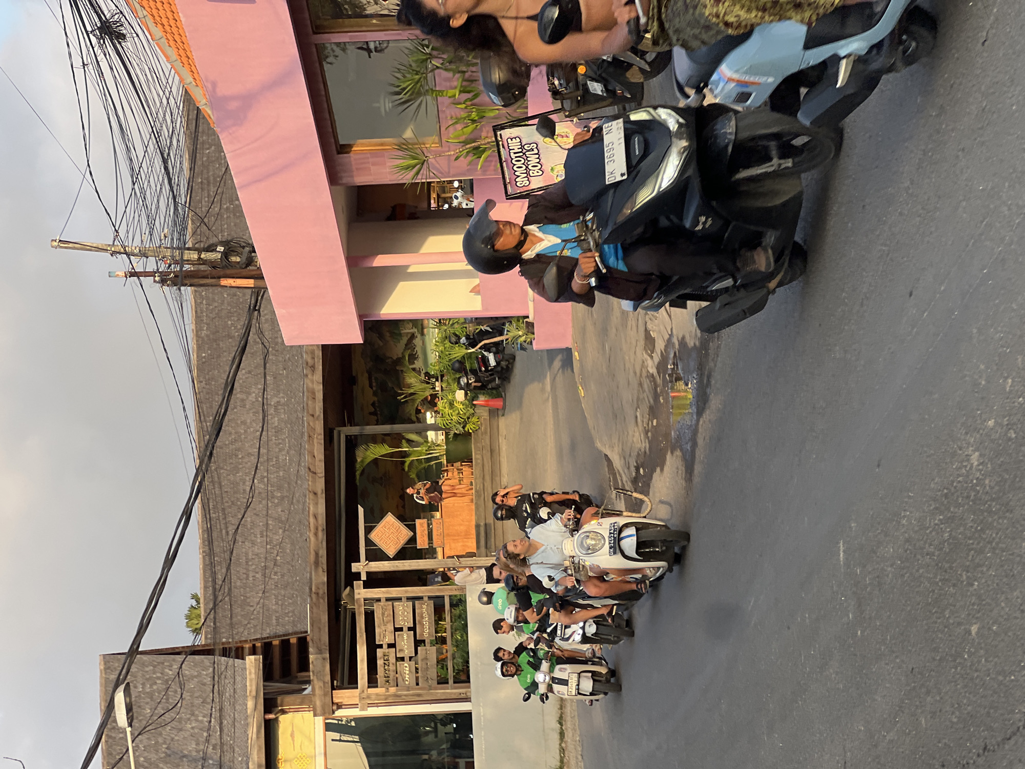 In Kuta Selatan, Indonesia, a group of people ride scooters down a street. The scene is illuminated by the golden hour sunlight, casting a warm glow on the buildings and road. Several scooters are in motion, with multiple individuals on each, some wearing helmets and casual attire. In the background, buildings with rustic wooden facades and a brightly colored pink structure suggest a commercial area, possibly with shops or eateries. A sign that reads "SMOOTHIE BOWLS" is visible, indicating a food service establishment. The street surface appears wet in patches, hinting at recent rain or cleaning. Overhead, a tangled network of power lines and poles traverses the sky, a common sight in urban Indonesian landscapes. The overall atmosphere is one of daily life and movement within a vibrant tropical setting.