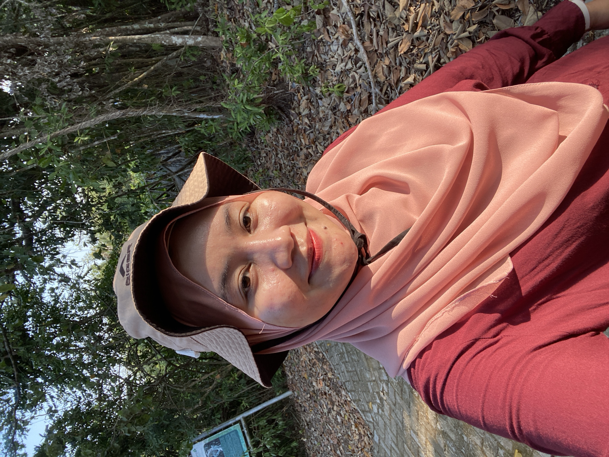 A medium shot shows a young woman wearing a light brown bucket hat and a pink hijab. She is also wearing a maroon long-sleeved shirt. She is smiling at the camera and her face is illuminated by sunlight. The background is a forest with trees and fallen leaves on the ground. There is a path on the left side of the image and a signpost with text and images. The location is the city of Sungai Pitung, Indonesia.