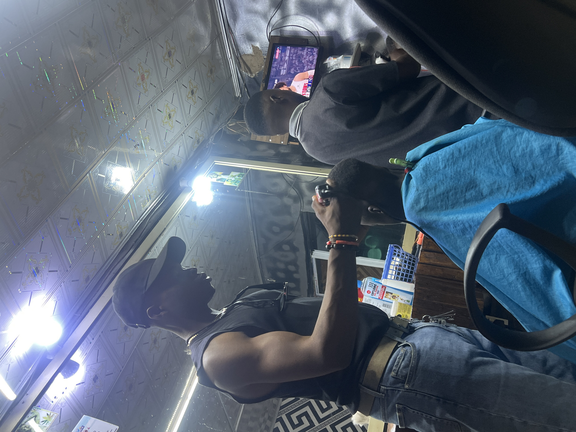 The image shows the interior of what appears to be a barbershop in Gura Top, Nigeria. A man wearing a black sleeveless shirt, blue jeans, a black cap, and bracelets on his right wrist is holding a device to his eye. He has a muscular build. A blue barber cape is partially visible on a seated customer to the right. Another man wearing a dark sweater sits in front of a small television mounted on the wall. The television displays a sports game. The walls and ceiling are covered in patterned, shiny, reflective panels. Bright artificial lights illuminate the scene. A shelf containing various products is visible in the background. The floor is patterned with a black and white geometric design.