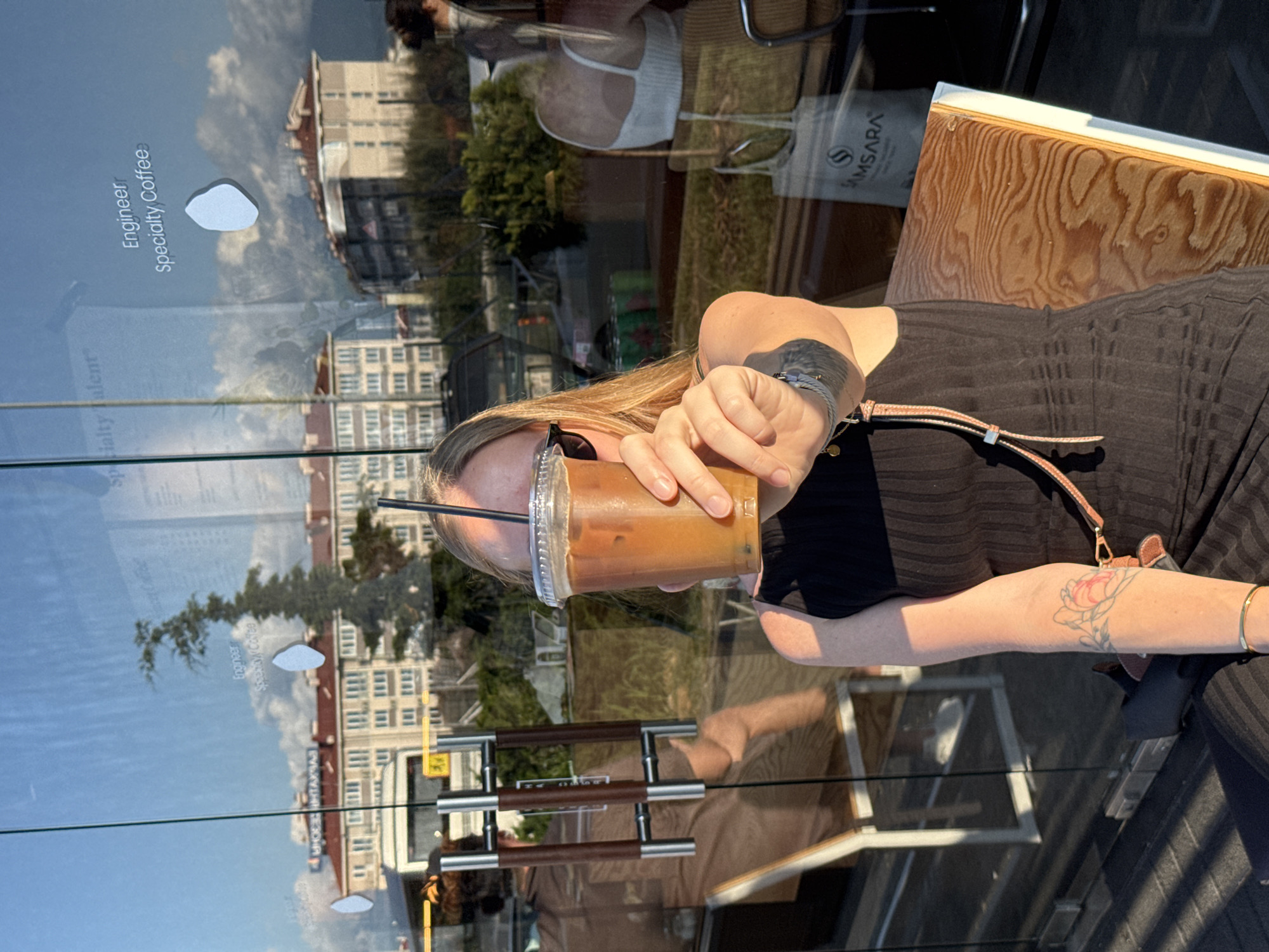 In Sochi, Russia, on a sunny day, a woman sits at an outdoor cafe, holding a plastic cup of iced coffee with a straw. She is wearing a black sleeveless dress and sunglasses. Reflected in the glass window of the cafe, we see buildings, trees, and clouds. The cafe's interior is also visible through the glass, showing tables, chairs, and other patrons. A sign on the window reads "Engineer Specialty Coffee."