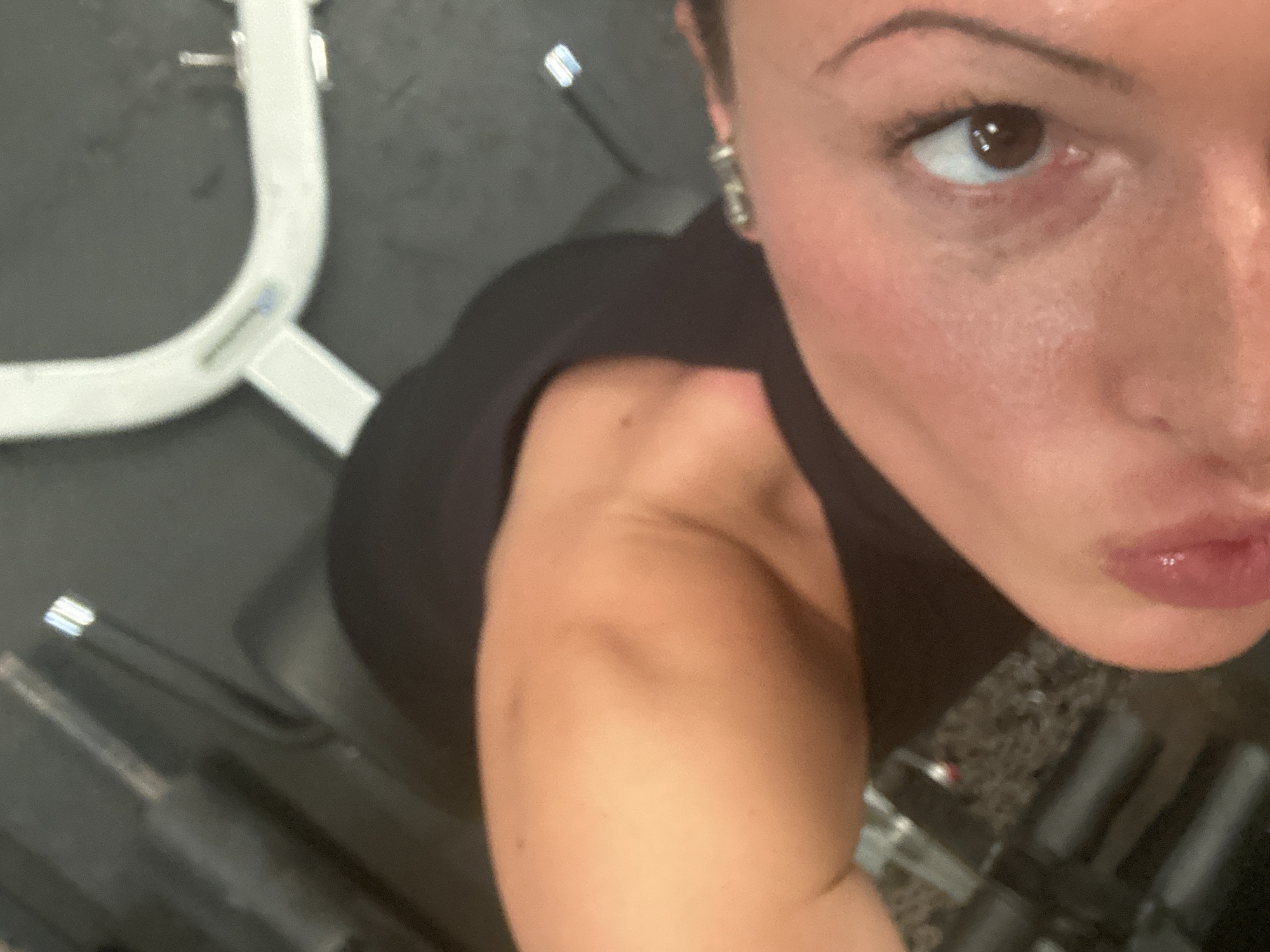 The image is a selfie taken from a high angle, focusing on the face and shoulder of a woman. She has brown eyes and is wearing a black top. The background reveals she is likely in a gym, with weightlifting equipment visible, including a bench press. There's no specific evidence to place the location in Tunbridge Wells, United Kingdom.