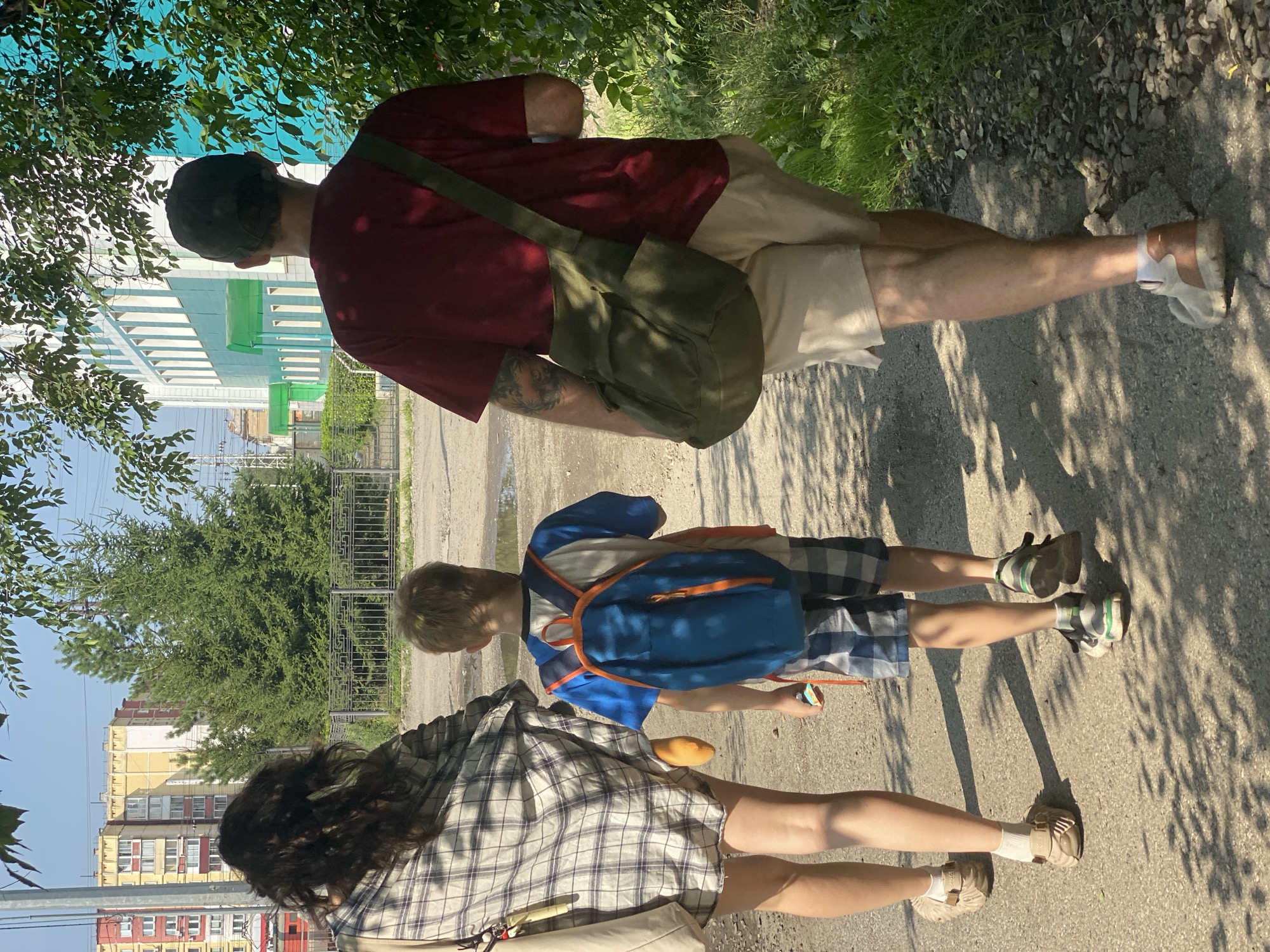 Three people, an adult male, an adult female, and a child, are walking away from the camera on a gravel path. The female, on the left, has long dark hair and is wearing a plaid shirt and tan slip-on shoes with white socks. She is carrying a light-colored bag over her shoulder. The child, in the center, is wearing a blue and orange backpack, a blue t-shirt, and plaid shorts. They are holding a small toy. The male, on the right, is wearing a dark red t-shirt, tan shorts, and light-colored sneakers. He has a green messenger bag slung over his shoulder and a baseball cap on. They are walking past a green building with white trim on the right and apartment buildings in the background on the left. Trees and foliage are visible on both sides of the path. The scene appears to be in Novokuznetsk, Russia, based on the architecture and urban setting. The lighting suggests it is a sunny day.