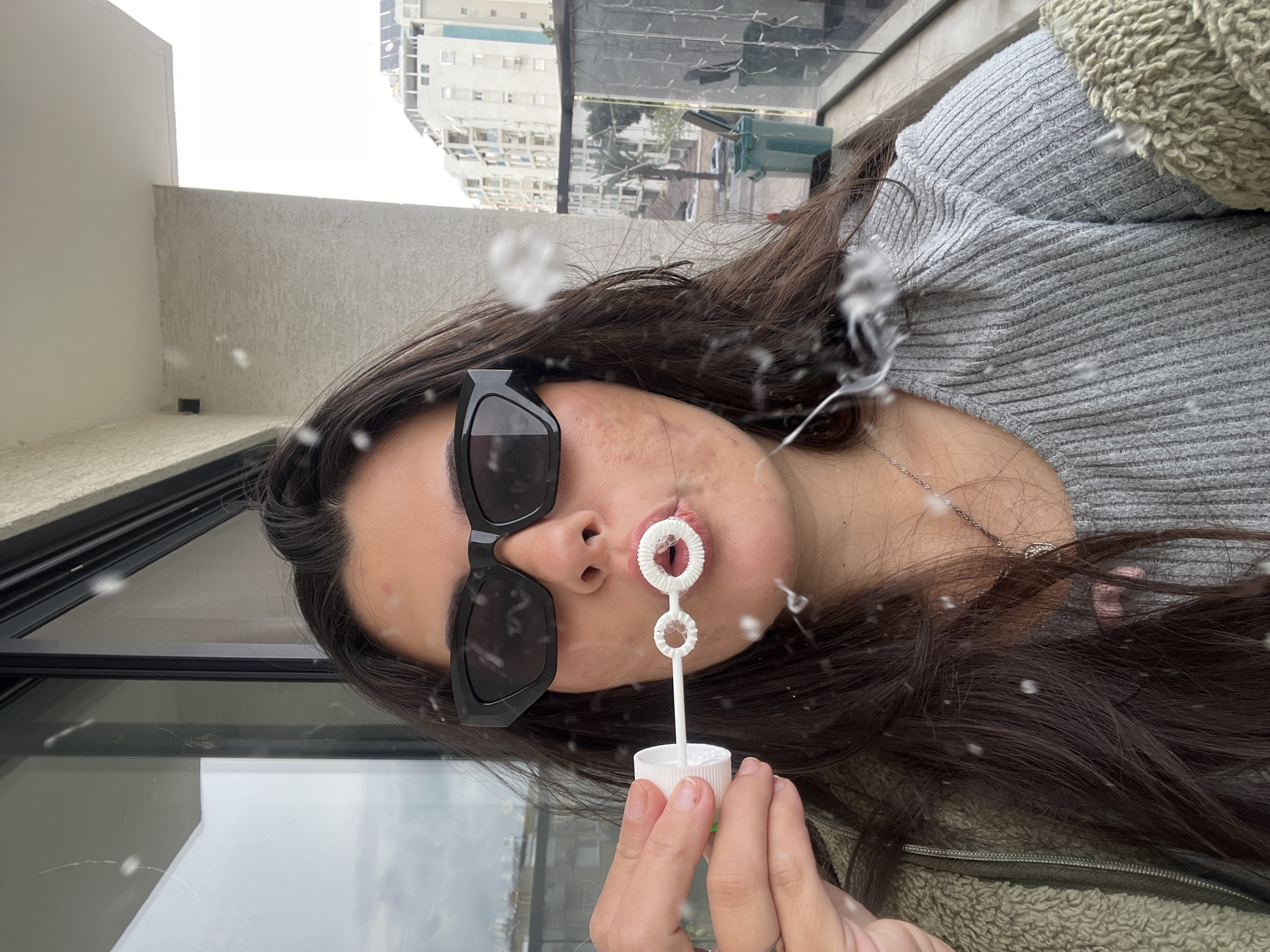 An adult female is depicted in a close-up, selfie-style photograph, standing on a residential balcony in Kiryat Bialik, Israel, on an overcast day. She has long, dark brown hair and wears black, angular sunglasses. Her attire consists of a light grey, ribbed long-sleeve top under an olive-green fuzzy jacket. She is actively blowing bubbles, with a white plastic bubble wand held to her pursed lips and the associated solution container in her left hand. Numerous small, foamy bubble fragments are suspended in the air around her face and hair.
The immediate background reveals a glass sliding door or window with a black frame to her left and a textured beige concrete balcony wall to her right. Above, a white concrete ceiling overhang is visible. Beyond the balcony's glass railing, a cityscape unfolds under a bright, cloudy sky. This includes multiple light-colored, multi-story buildings, some featuring rooftop solar panels or water heaters. A street is visible below with a prominent palm tree, at least one parked vehicle, and two green garbage bins.