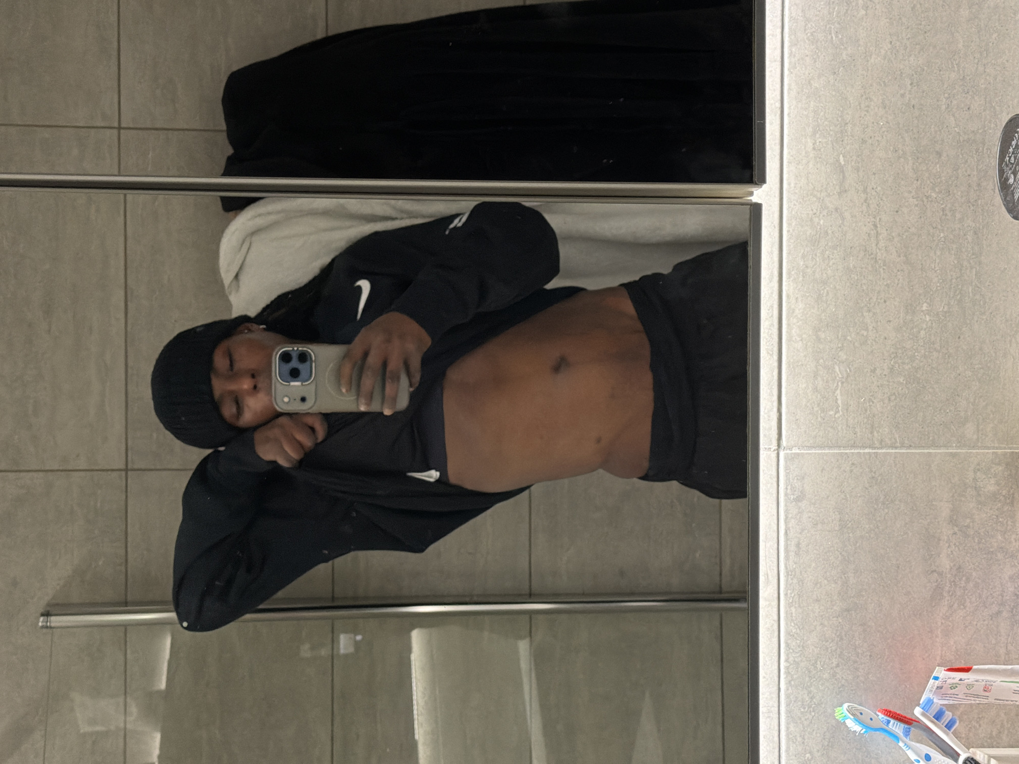 A person is taking a mirror selfie in a bathroom. The person is wearing a black beanie, a black long-sleeve shirt with a white Nike swoosh logo, and black pants or shorts. The person is holding an iPhone in their right hand, partially obscuring their face. The person's torso is exposed, revealing some abdominal definition.
The bathroom has light grey tiled walls. A black towel is hanging above the person in the mirror. A silver towel bar is visible near the bottom of the mirror. A shower enclosure with a partially visible glass door is also present. In the lower right corner of the image, two toothbrushes and a tube of toothpaste are visible.
There is no information available to determine if this location is in London, United Kingdom.
