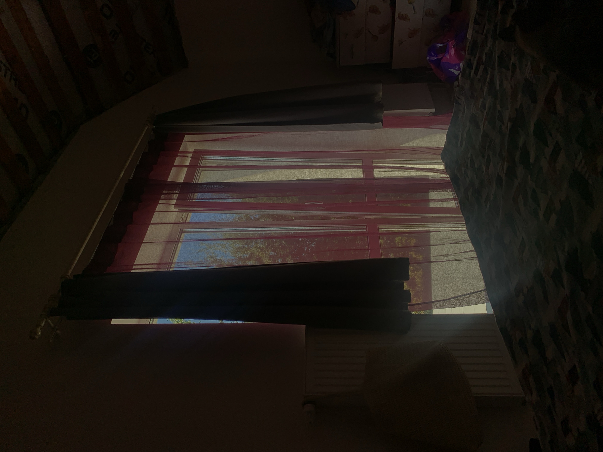 A dimly lit bedroom is captured, with natural light streaming in from a large window. The window is covered by two sets of curtains: dark, opaque curtains on the sides and sheer pink curtains in the center. Through the sheer curtains, trees and the sky are visible, suggesting an outdoor environment beyond the window. The light from the window casts a diagonal ray across a bed in the foreground, which has a patterned comforter. To the right of the bed, a chest of drawers is partially visible, with some items on top. The ceiling of the room appears to be slanted and made of wood. The overall impression is of a domestic setting, likely a bedroom, with the time of day suggested by the quality of the light being either early morning or late afternoon. This scene does not depict any specific location within the city of Minsk, Belarus, nor does it show any people or actions.