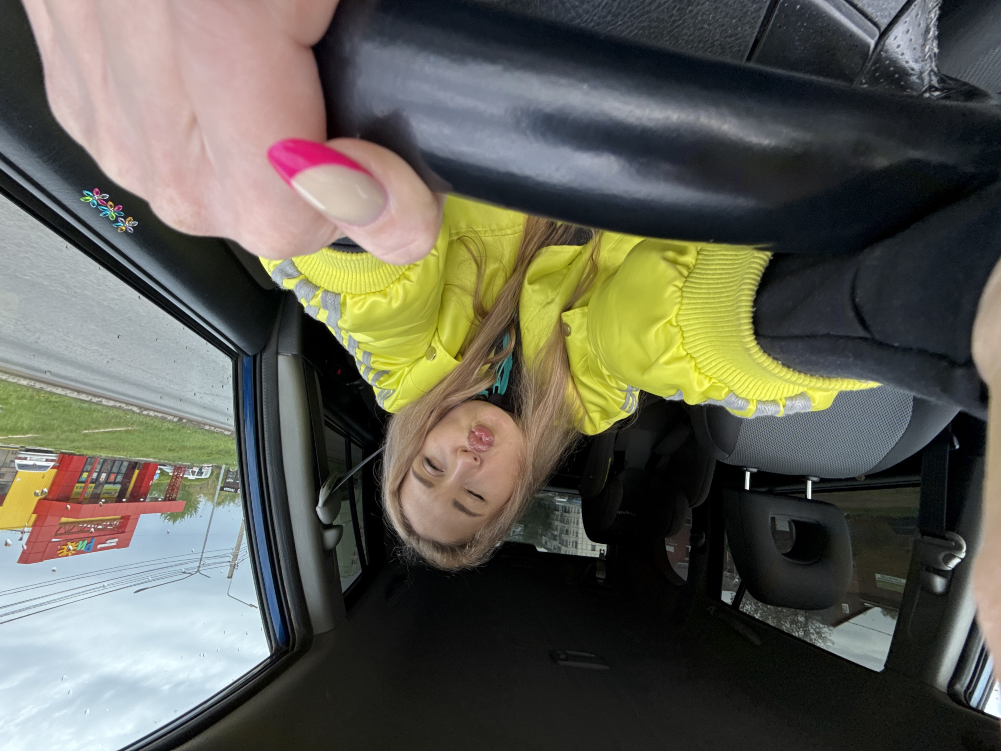 A blonde woman in a yellow jacket sits in the driver's seat of a car, upside down in the frame, holding the steering wheel. A pink-tipped nail is visible on her hand. Outside the car window is a building with a yellow and red facade. A sign on the building reads "ПЕЧИ". The image suggests a location in Chaikovsky, Russia, given the Russian signage. A child's seat is visible in the back.