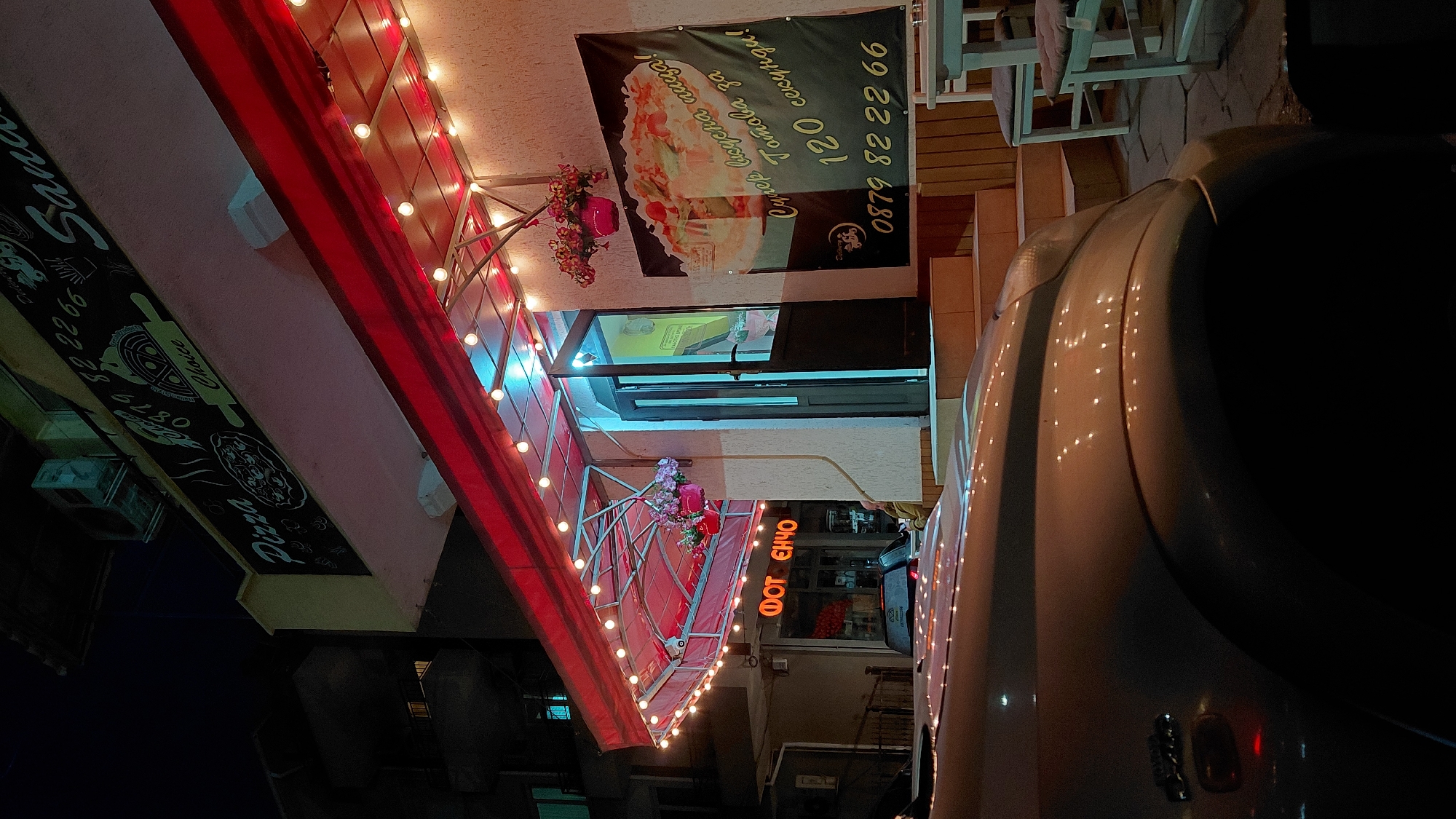 Here is a description of the image:
The image shows a nighttime scene of a storefront in an urban setting. The word "Pizza" is visible on a sign on the left side of the frame, along with a phone number. Above the entrance, a sign advertises "Супер вкусна пица! Готова за 120 секунди!" (Super delicious pizza! Ready in 120 seconds!). The number "0879 82 22 66" is also displayed on the sign.
The entrance to the store is flanked by decorations, including flowers and lights. In the lower right, a portion of a vehicle is visible, obscuring part of the scene. Additionally, a sign with the Bulgarian text "ФОТ ЕНЧО" (FOT ENCHO) can be seen.