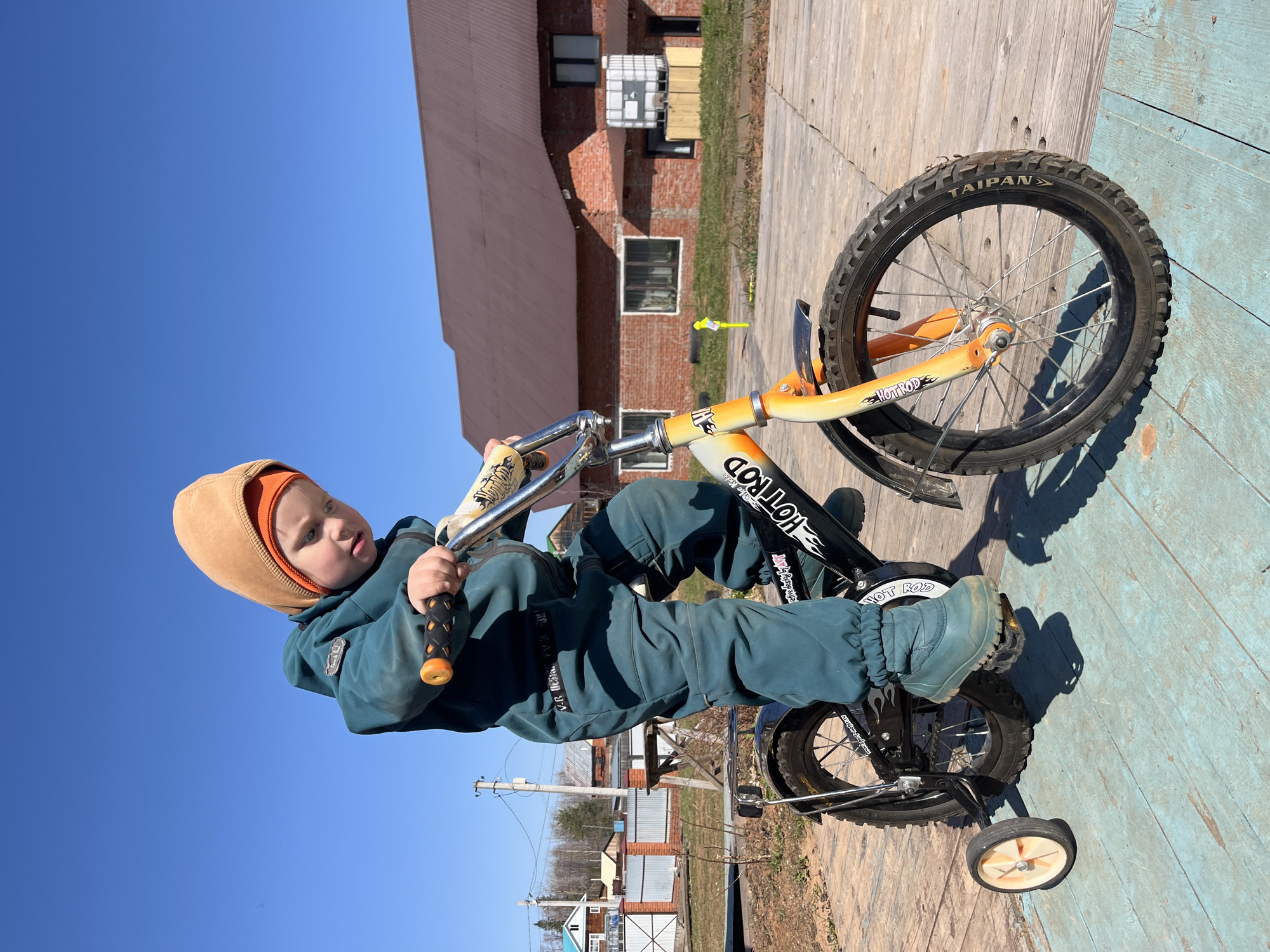 A young child in a teal jumpsuit and a brown hat is sitting on a small bicycle with training wheels on a light blue wooden deck. The bicycle has black and orange accents with the words "HOT ROD" printed on the frame. The front tire reads "TAIPAN." The child is looking to the left of the frame and holding the handlebars. In the background, there is a red brick building and a clear blue sky. The location appears to be outdoors in the city of Chaikovsky, Russia, given the context. There are other buildings and utility poles visible in the distance. The scene is well-lit.