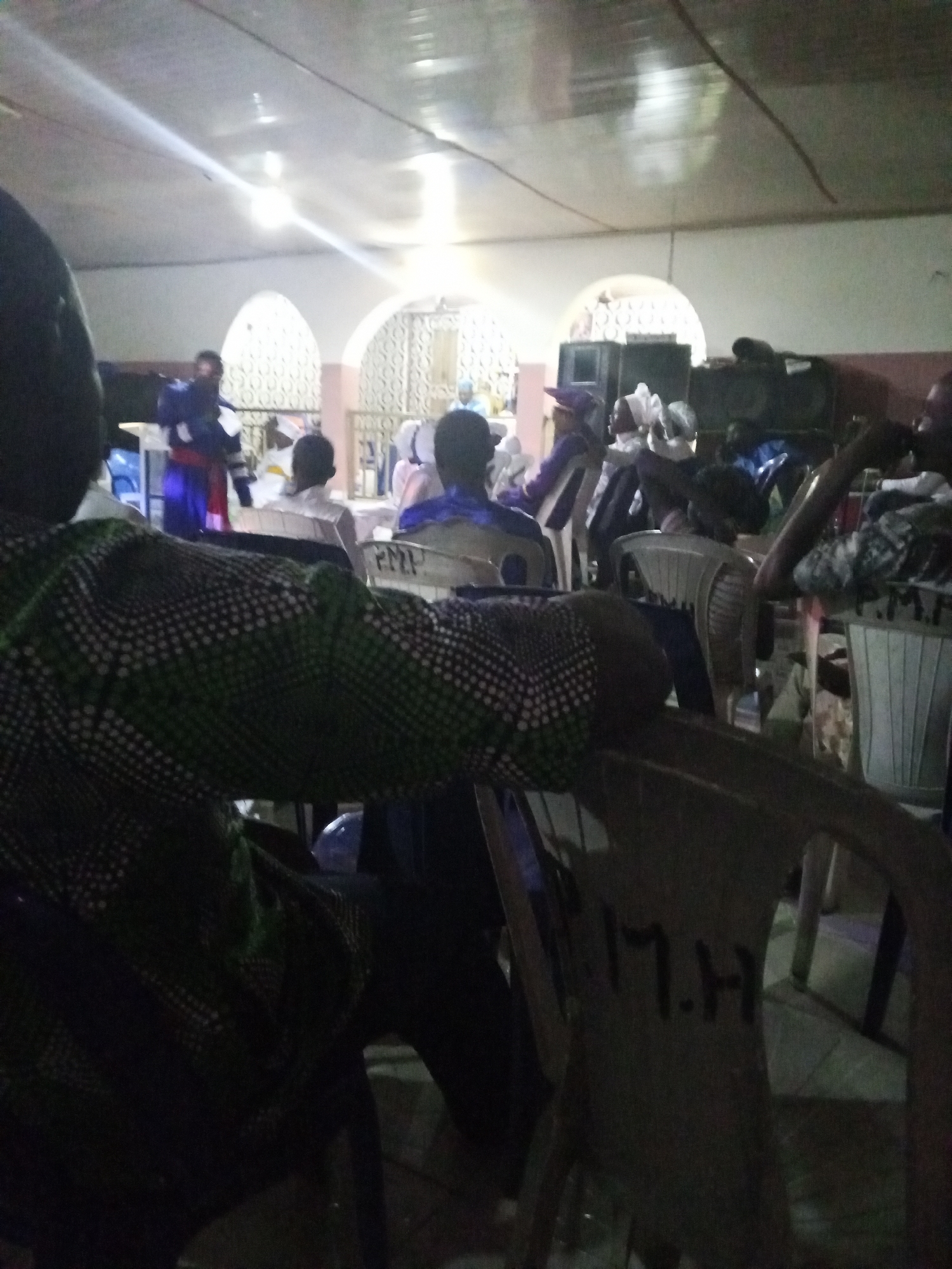 The image appears to show a gathering of people indoors, likely in a church or meeting hall. The location is possibly in Lokoja, Nigeria, though this cannot be definitively confirmed from the image alone. The lighting is dim, and the focus is somewhat blurry.
Several individuals are seated on plastic chairs, facing towards a raised platform where one person is standing and possibly speaking or performing. The person on the platform is wearing a distinctive blue and red garment. The seated individuals seem to be an audience.
The room is simple, with arches in the background and what appears to be audio equipment. The overall atmosphere is that of a religious service or a community meeting.