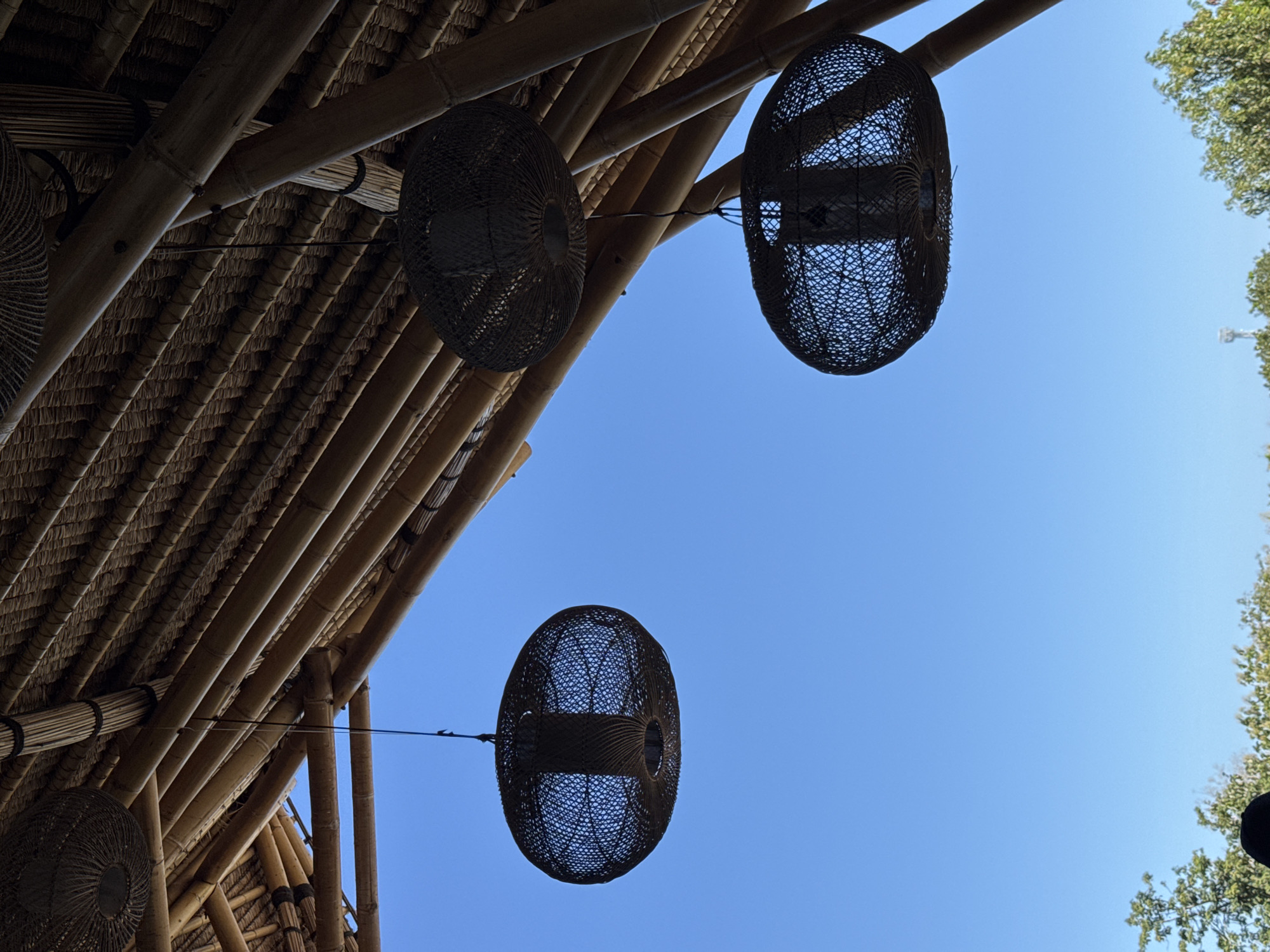 The image, likely taken in Kuta Selatan, Indonesia, presents a low-angle view of a bamboo structure with hanging woven lamps against a clear blue sky. The structure features a bamboo frame supporting a thatched roof, partially visible in the upper left. Three woven lamps, spherical in shape, are suspended from the structure by wires, positioned against the unobstructed blue sky. The top right edge of the image shows green tree foliage against the same blue sky.